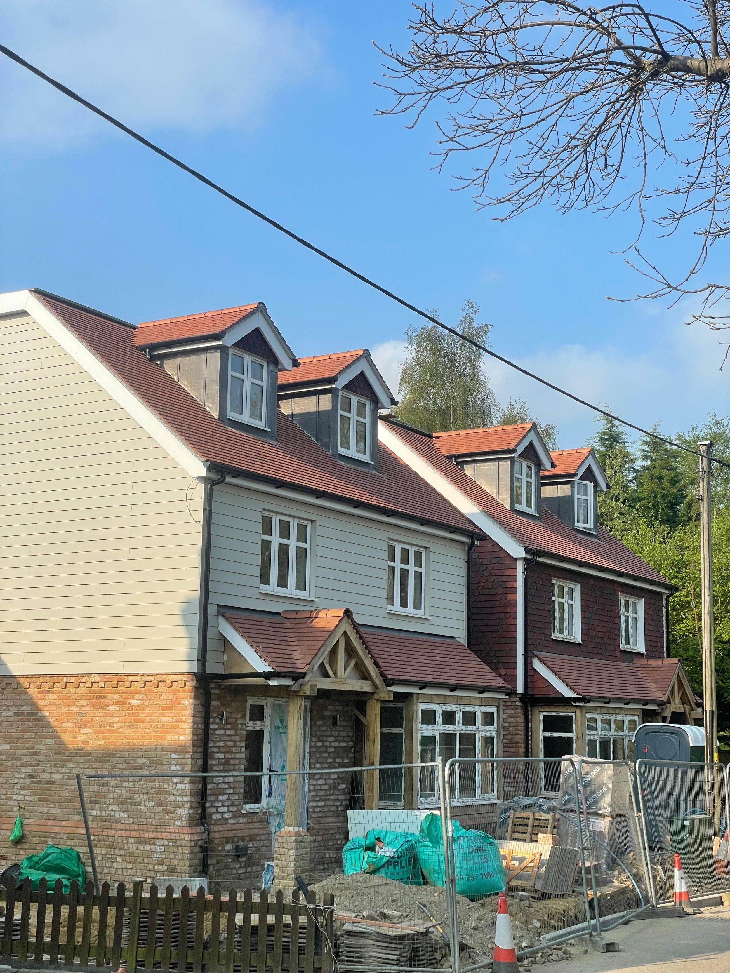 Construction site in front of a brick row house with beige and dark red siding and red tile roof, multiple dormer windows, and orange construction cones.