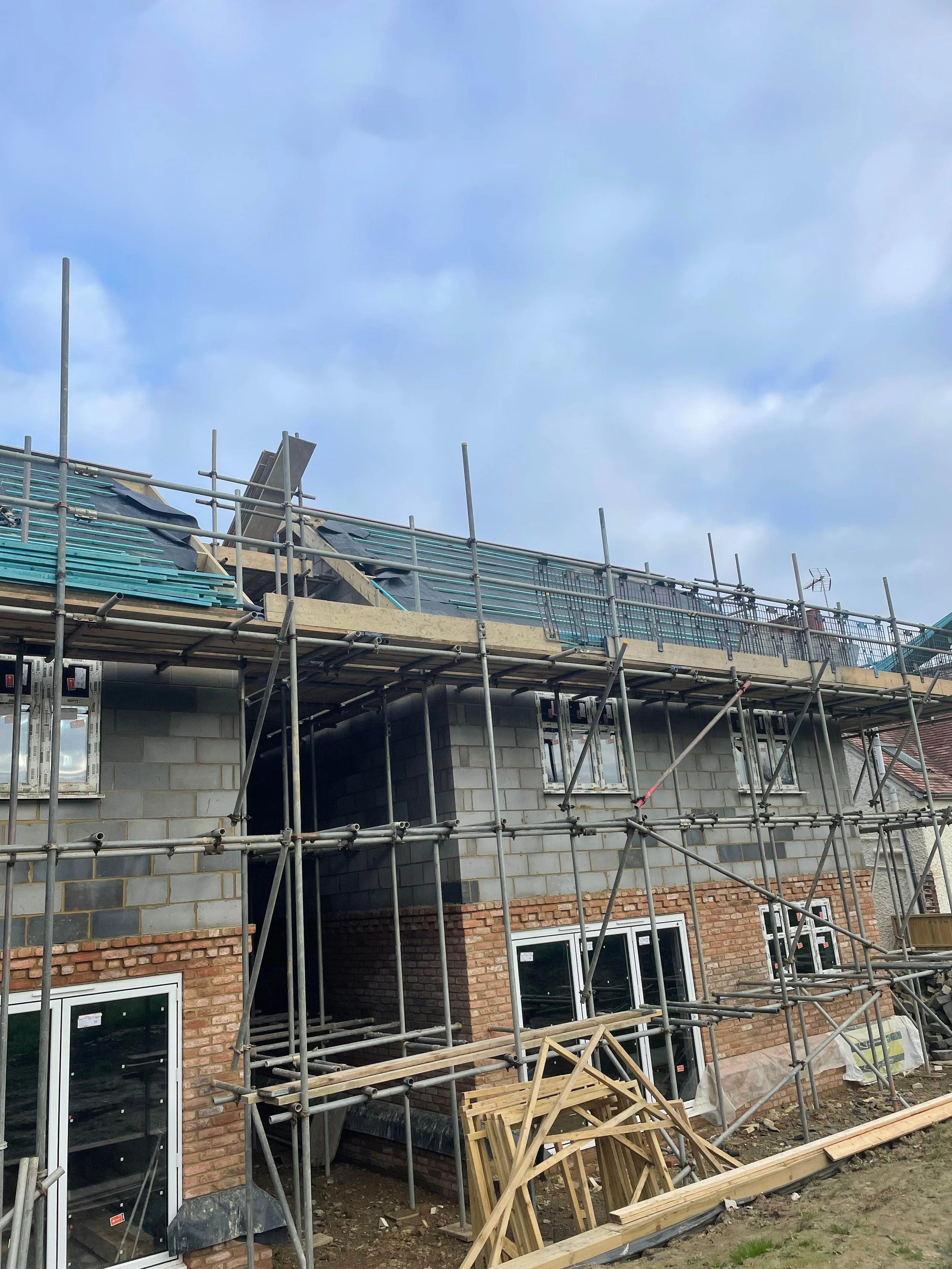 Under construction house with scaffolding surrounding the building, brick and concrete walls, and some construction materials on the ground; cloudy sky overhead.