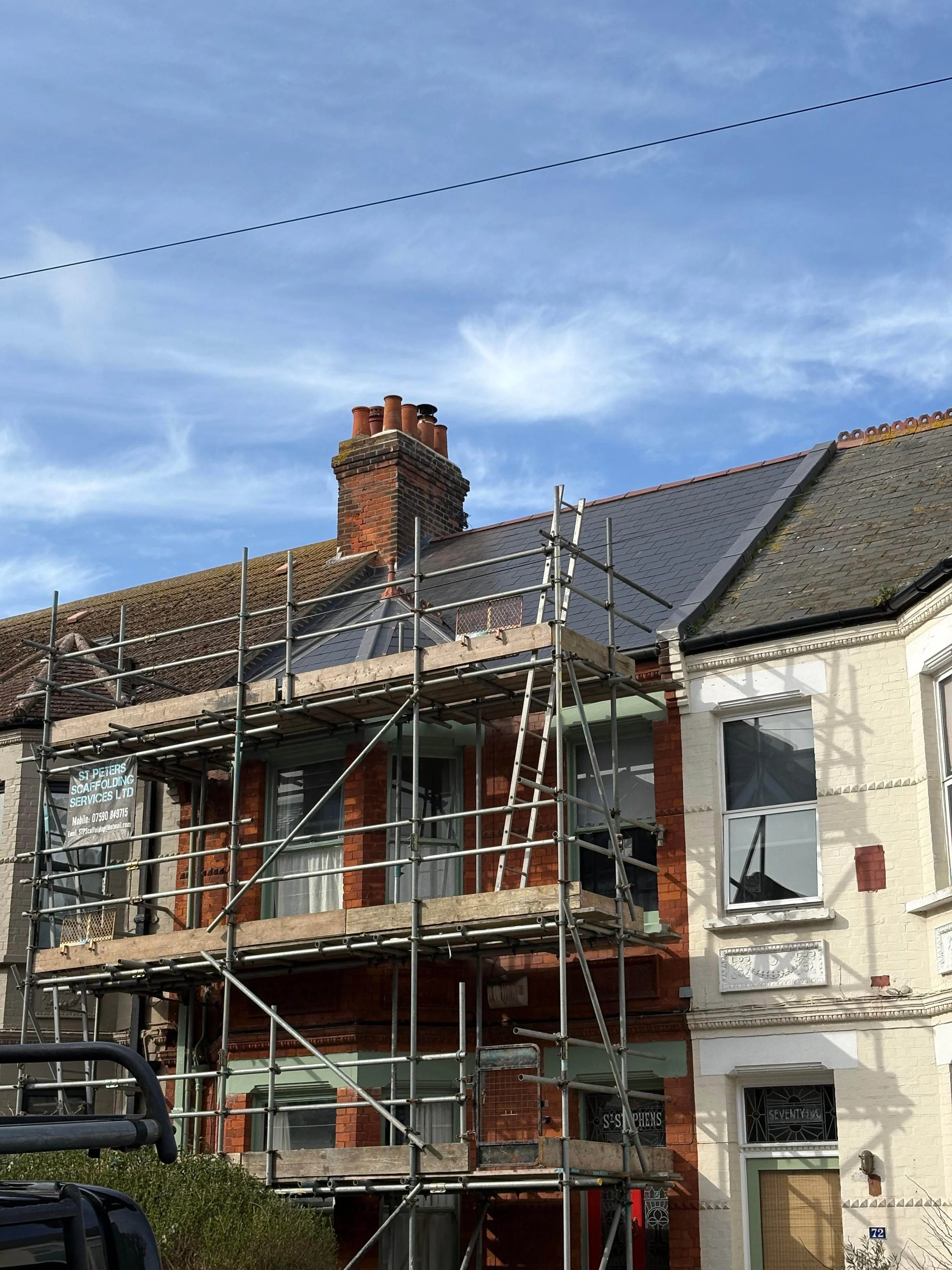 A house under renovation with scaffolding on the front, partially covered with wooden planks. The house has a brick exterior and two large windows with curtains. The neighboring house on the right is painted white. The sky is mostly clear with some w
