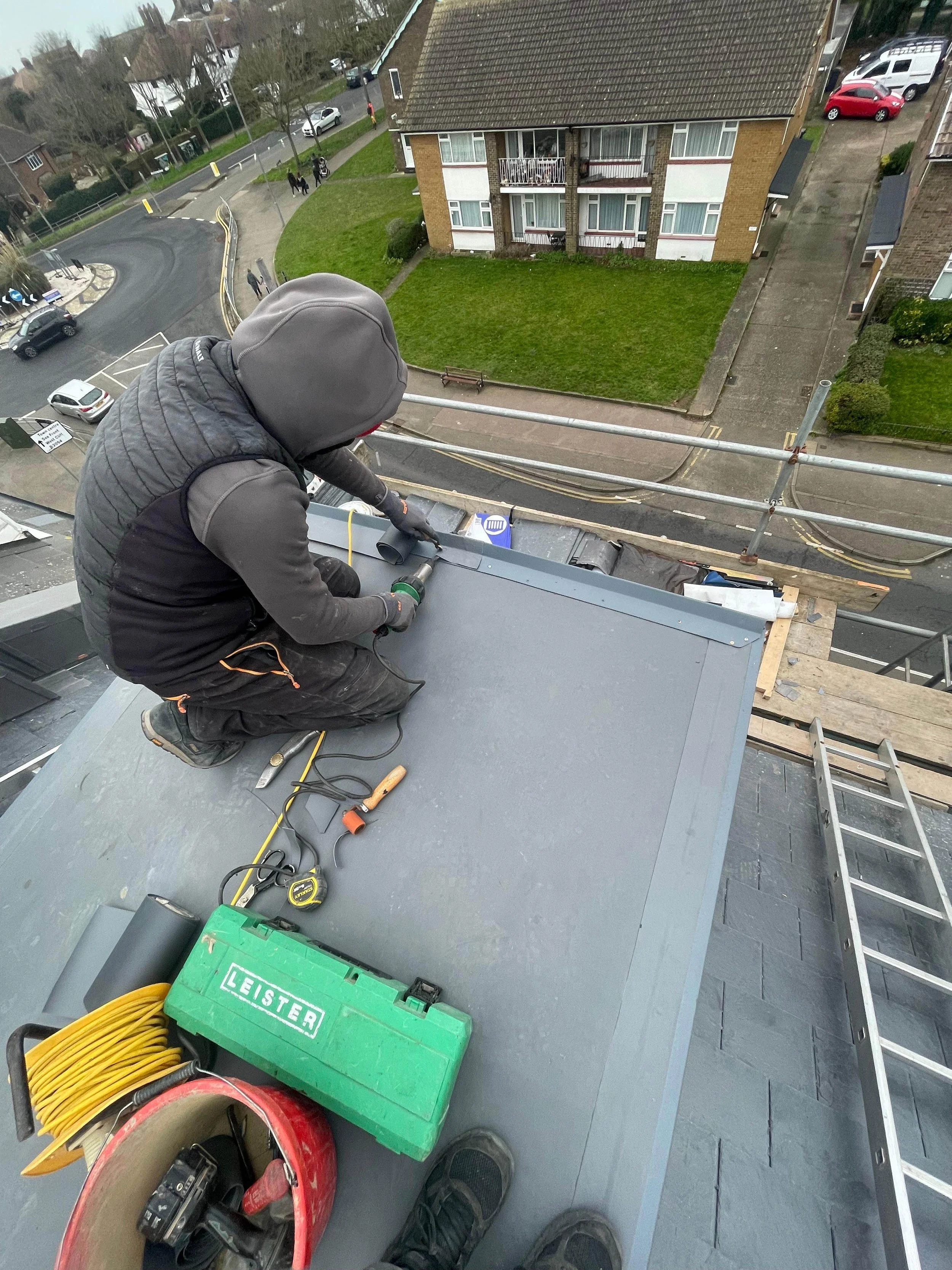 A worker on a roof using a drill or screwdriver to work on the roof of a building, with construction tools and equipment nearby, including a green box labeled 'LEISTER', a tape measure, a cordless drill, a roll of tape, extension cords, and a ladder.