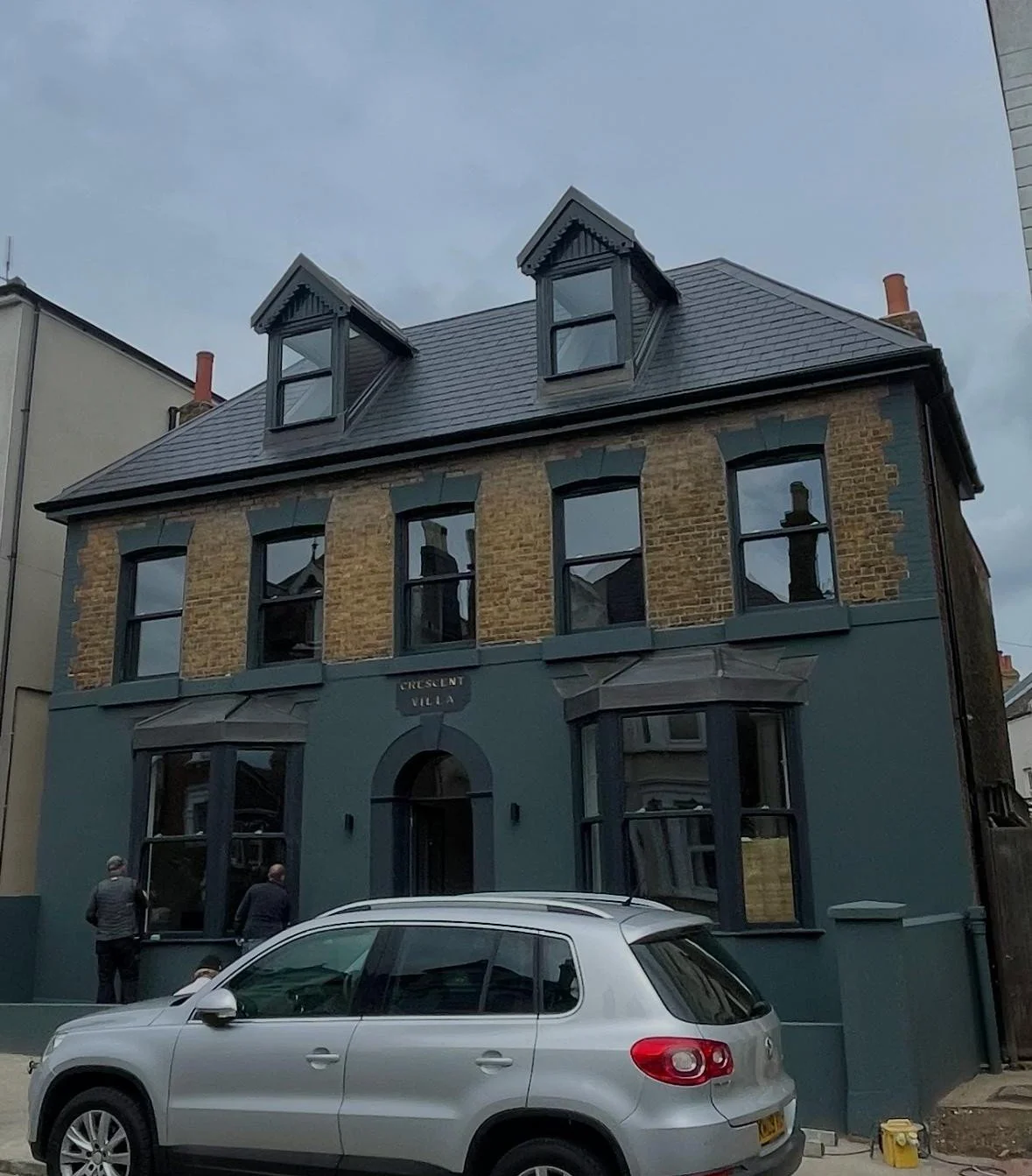 A three-story building with a blue painted facade, large front windows, and a brick upper level with tall, narrow windows. Two small dormer windows are on the roof, and there are two people walking near the entrance. A silver car is parked in front o