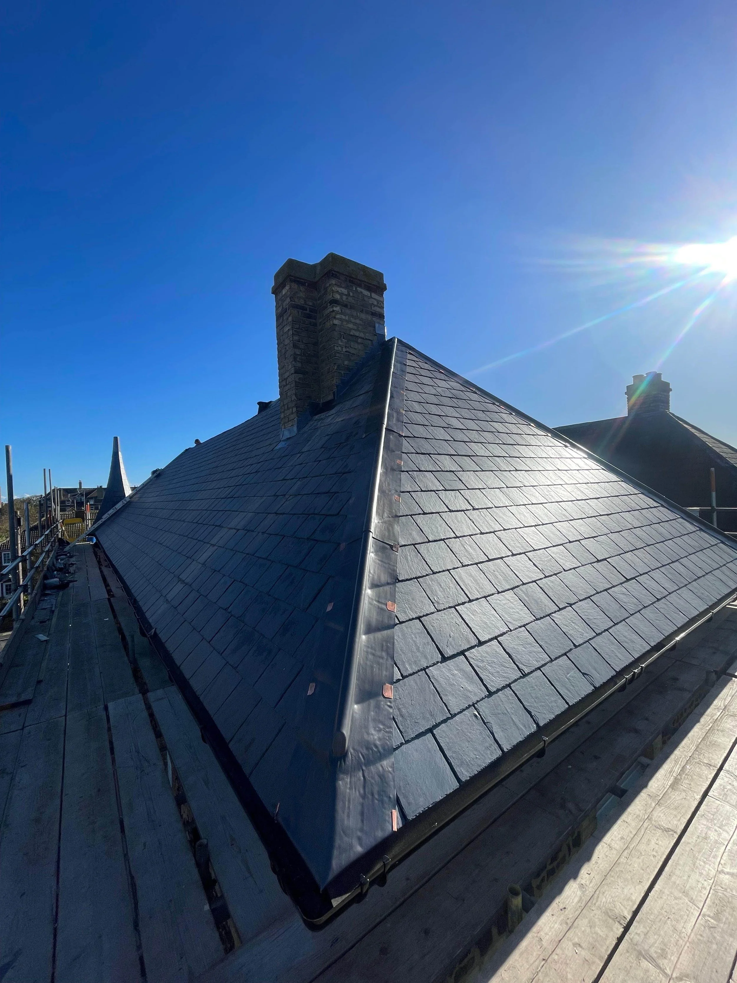 A rooftop with slate shingles and brick chimneys, under a clear blue sky with the sun shining brightly.
