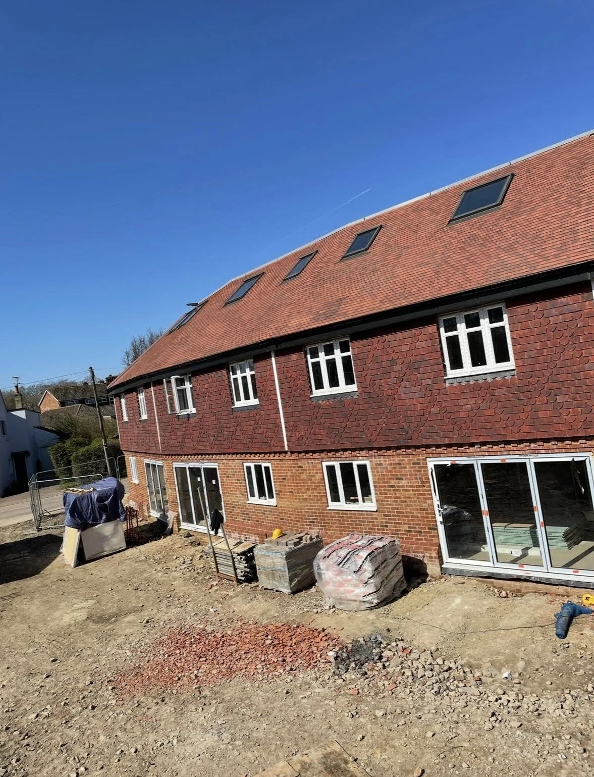 A multi-story house under construction with a red brick exterior, white-framed windows, and a sloped roof with skylights. The yard is unfinished with construction materials and debris.
