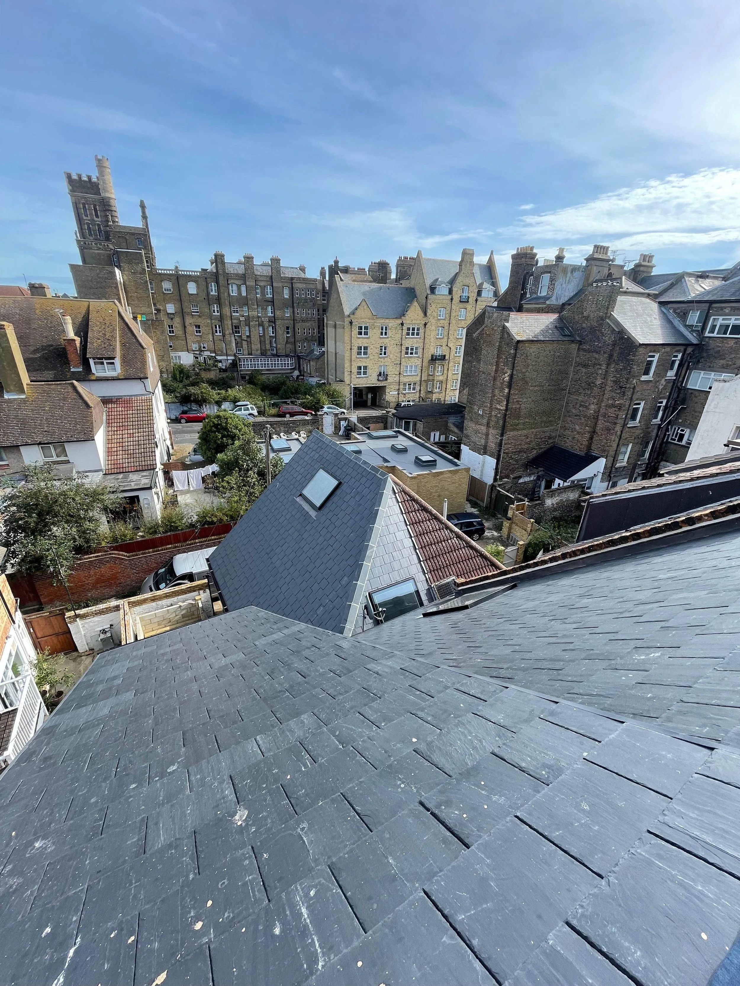 View from a rooftop showing neighboring roofs, trees, cars, and historic buildings under a blue sky with clouds.