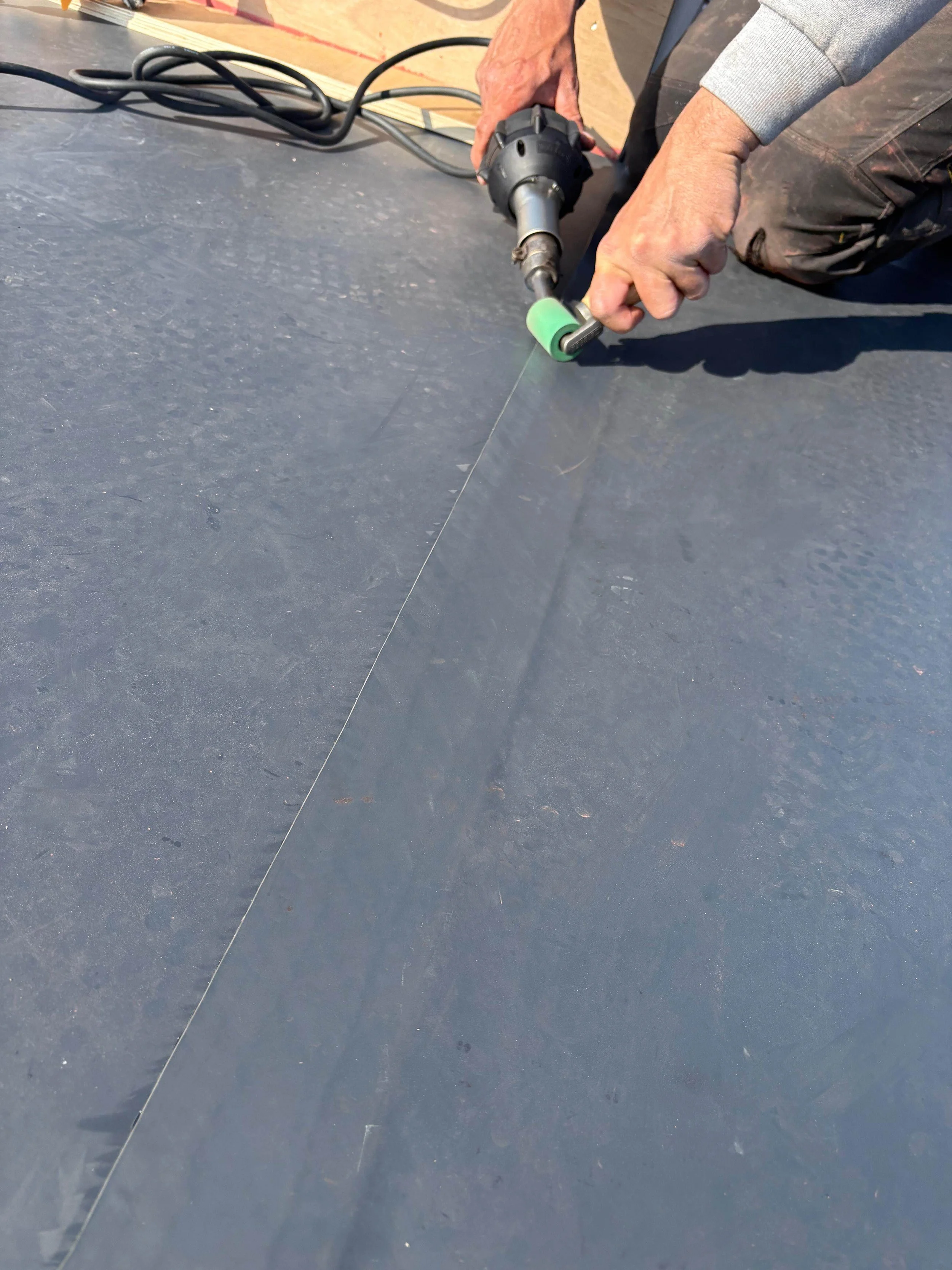 A person using a power tool to cut a straight line on a dark, textured surface, possibly flooring or roofing.