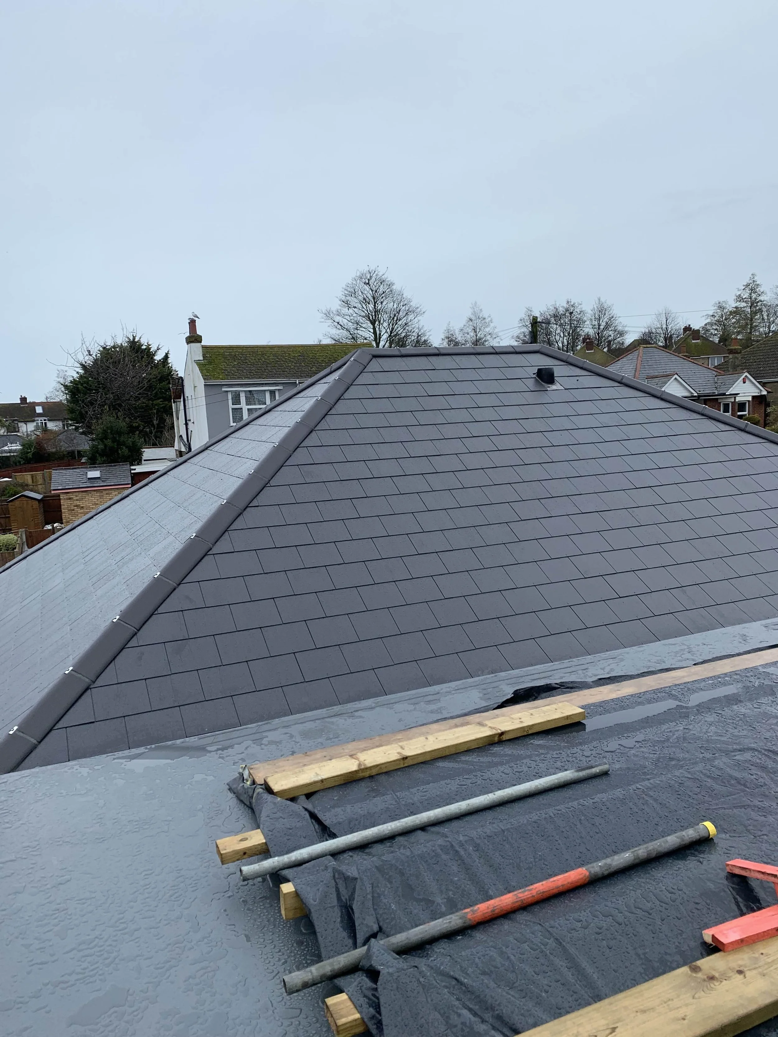View of a newly installed gray tiled roof on a house with some construction tools and materials on the foreground and neighboring houses in the background under a cloudy sky.