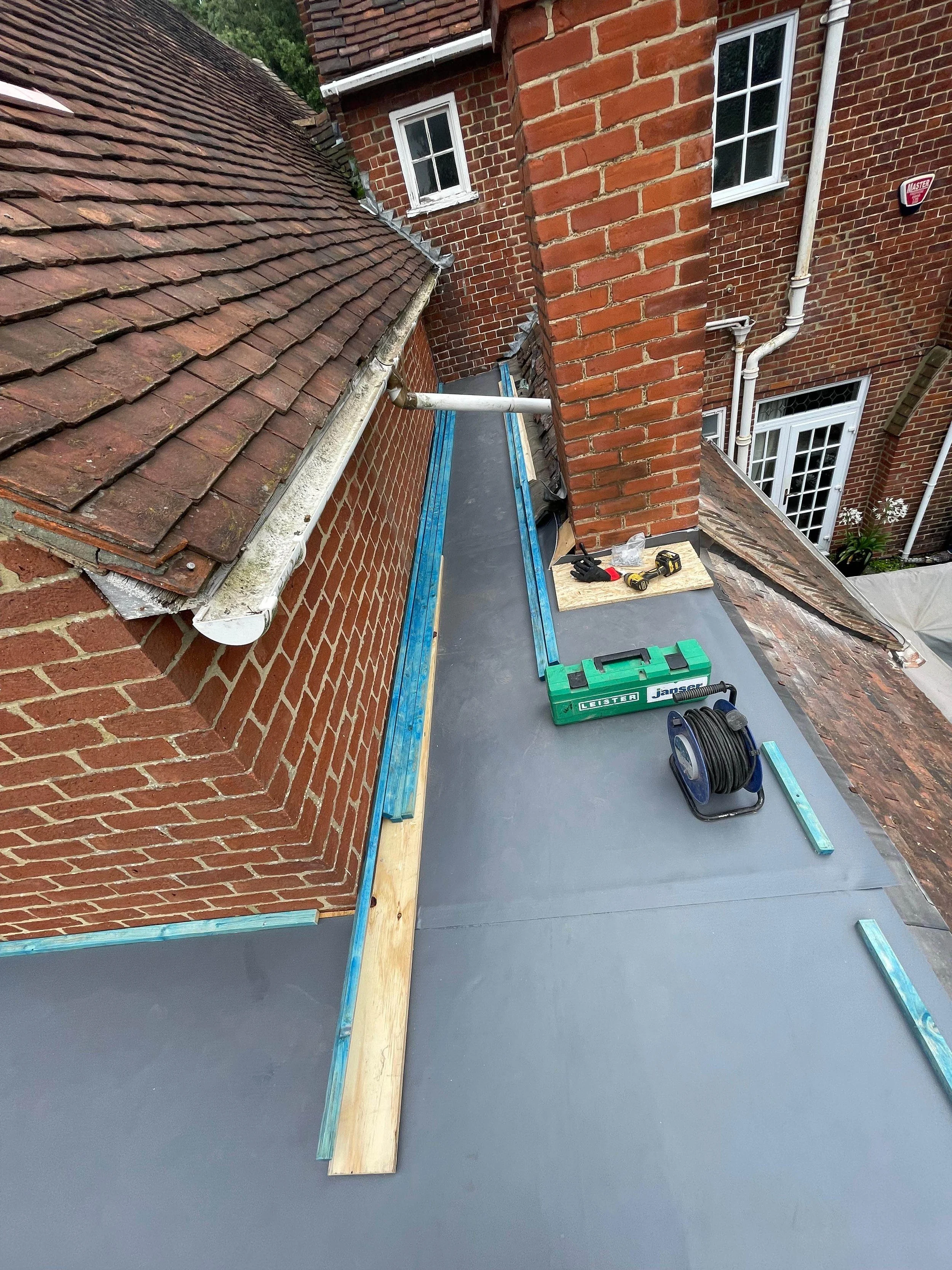 Roof construction work with tools, wooden beams, and weatherproofing materials on a residential building with brick exterior.