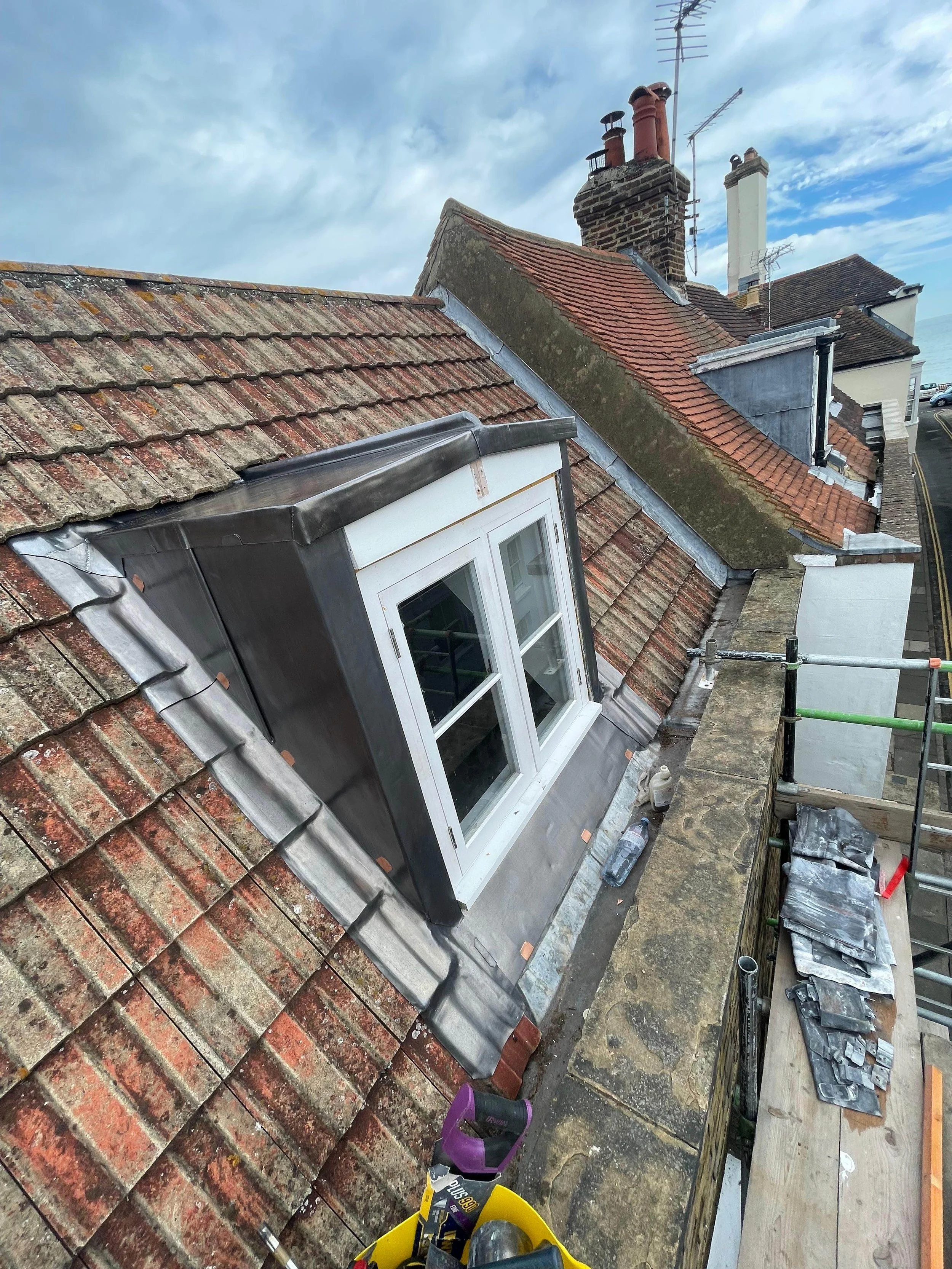 Roof undergoing renovation with a new dormer window installed, construction tools and materials on scaffolding, chimney stacks visible, and partly cloudy sky.