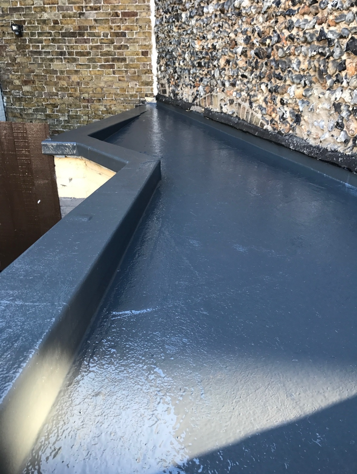 A newly painted black flat rooftop with a small raised parapet wall along the edge, adjacent to a brick wall and a pebbled surface.