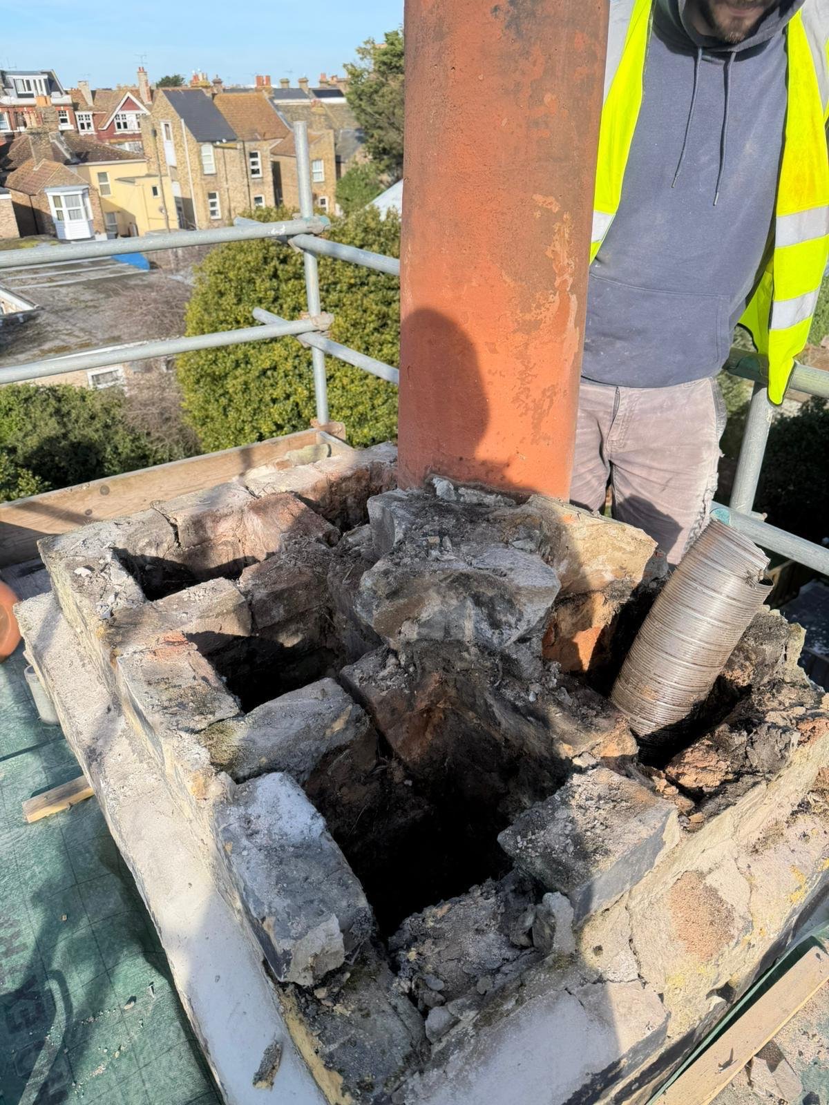 Close-up of a chimney repair site on a building's rooftop showing exposed bricks, a new metal pipe, and a worker in a yellow safety vest and gray hoodie.