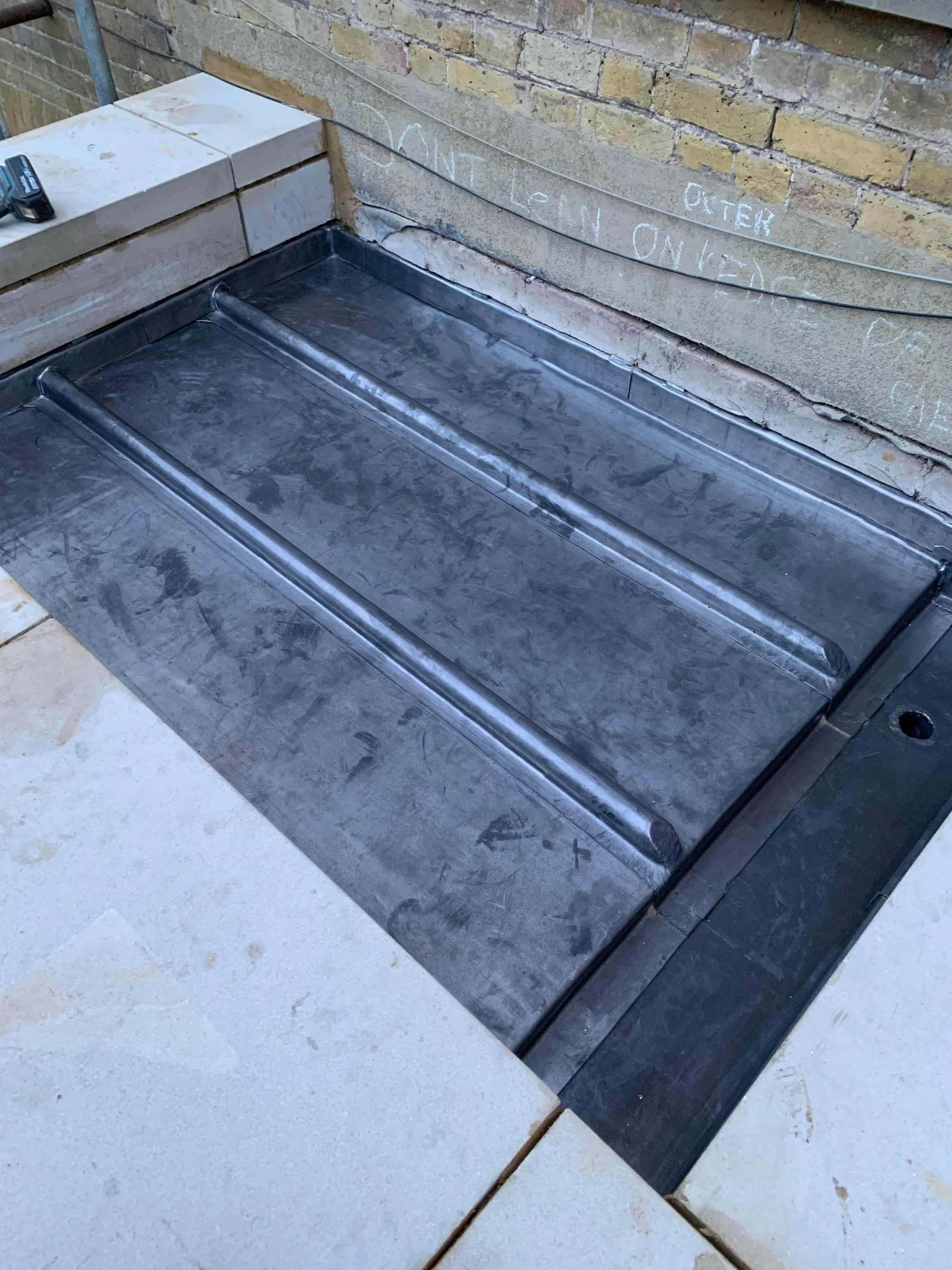 A metal fire pit with a rectangular shape, black frame, and three raised ridges on the surface, installed on a brick and tile patio with a brick wall in the background.