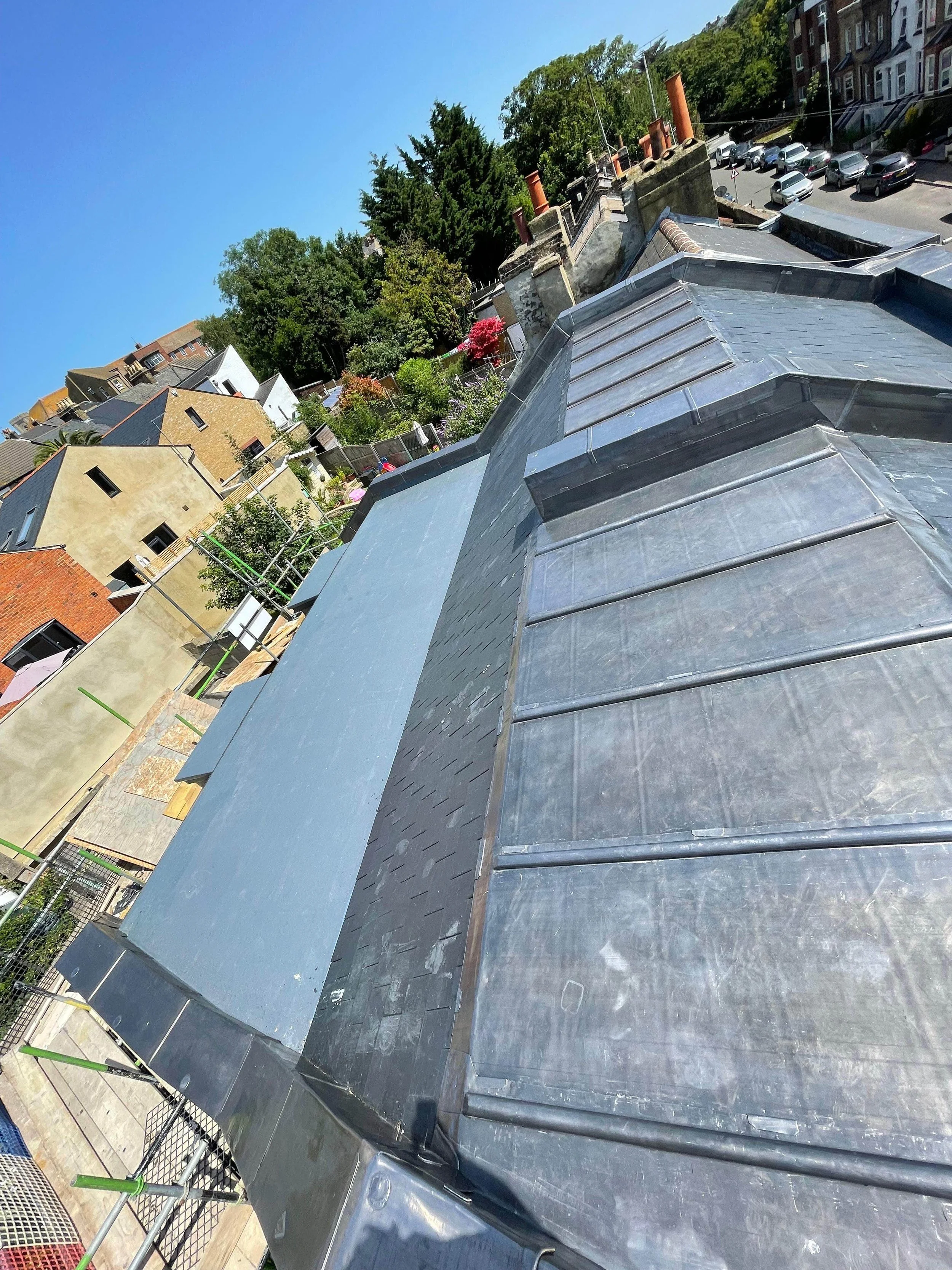 View of a rooftop with metal roofing, neighboring houses, trees, and a street with cars parked along it under a clear blue sky.