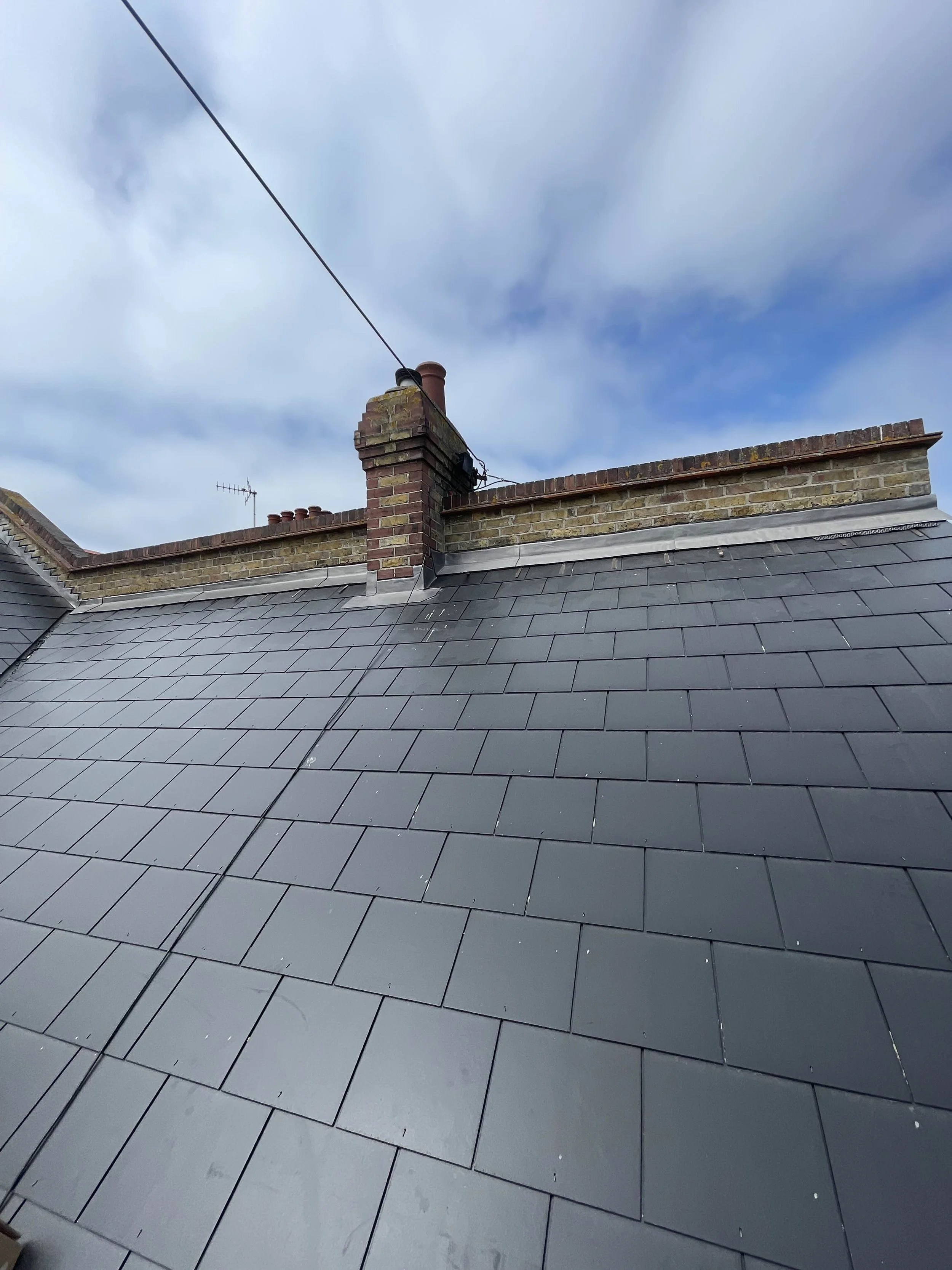 View of a brick chimney and roof with black slate tiles on a building under a cloudy sky.