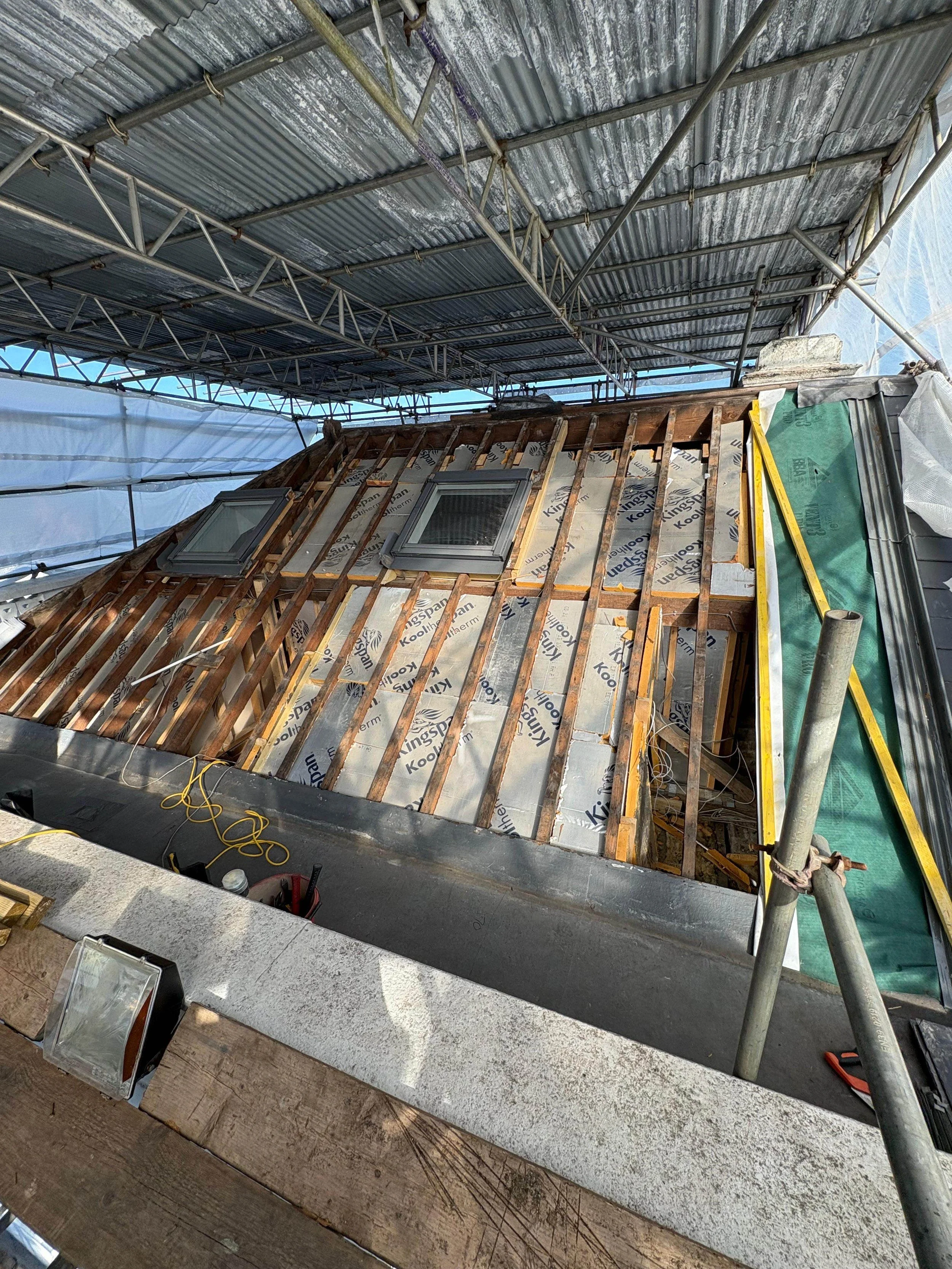 Construction site on a roof with an incomplete dormer window, exposed wooden framing, insulation, and roofing materials. Scaffolding surrounds the area.