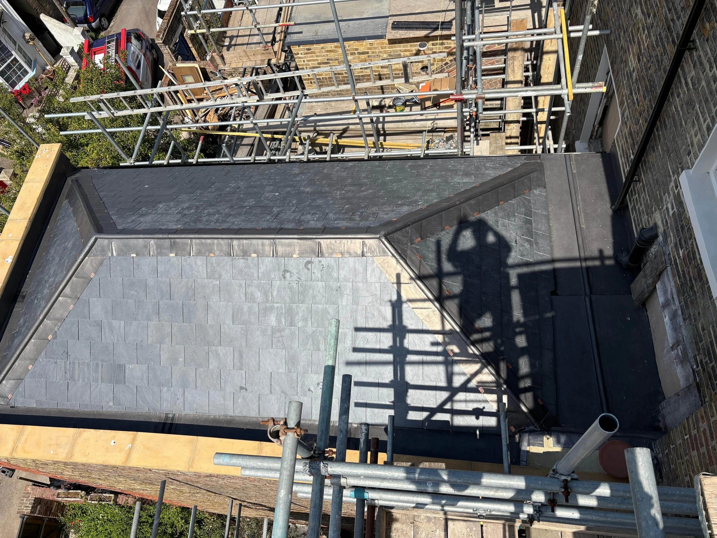 View of a rooftop with grey tiles, some scaffolding around the edges, and construction materials. There is a shadow of a person taking the photo. In the background, there are neighboring buildings, trees, and cars visible.