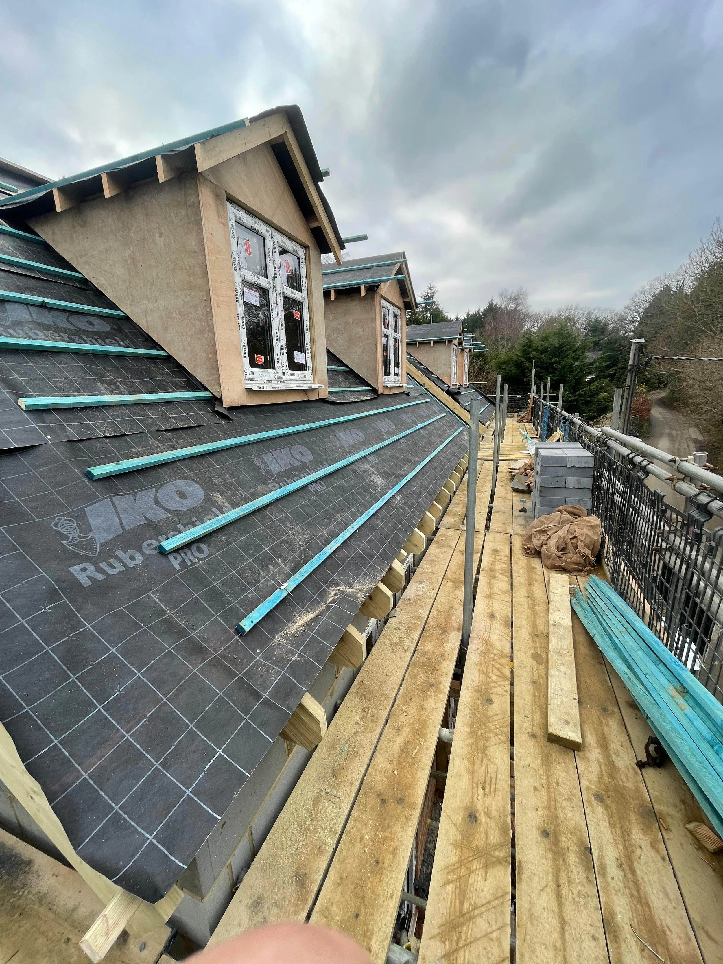 Under construction on a roof with black roofing underlayment, dormer windows, and wooden framing, with a safety walkway along the edge and trees in the background.