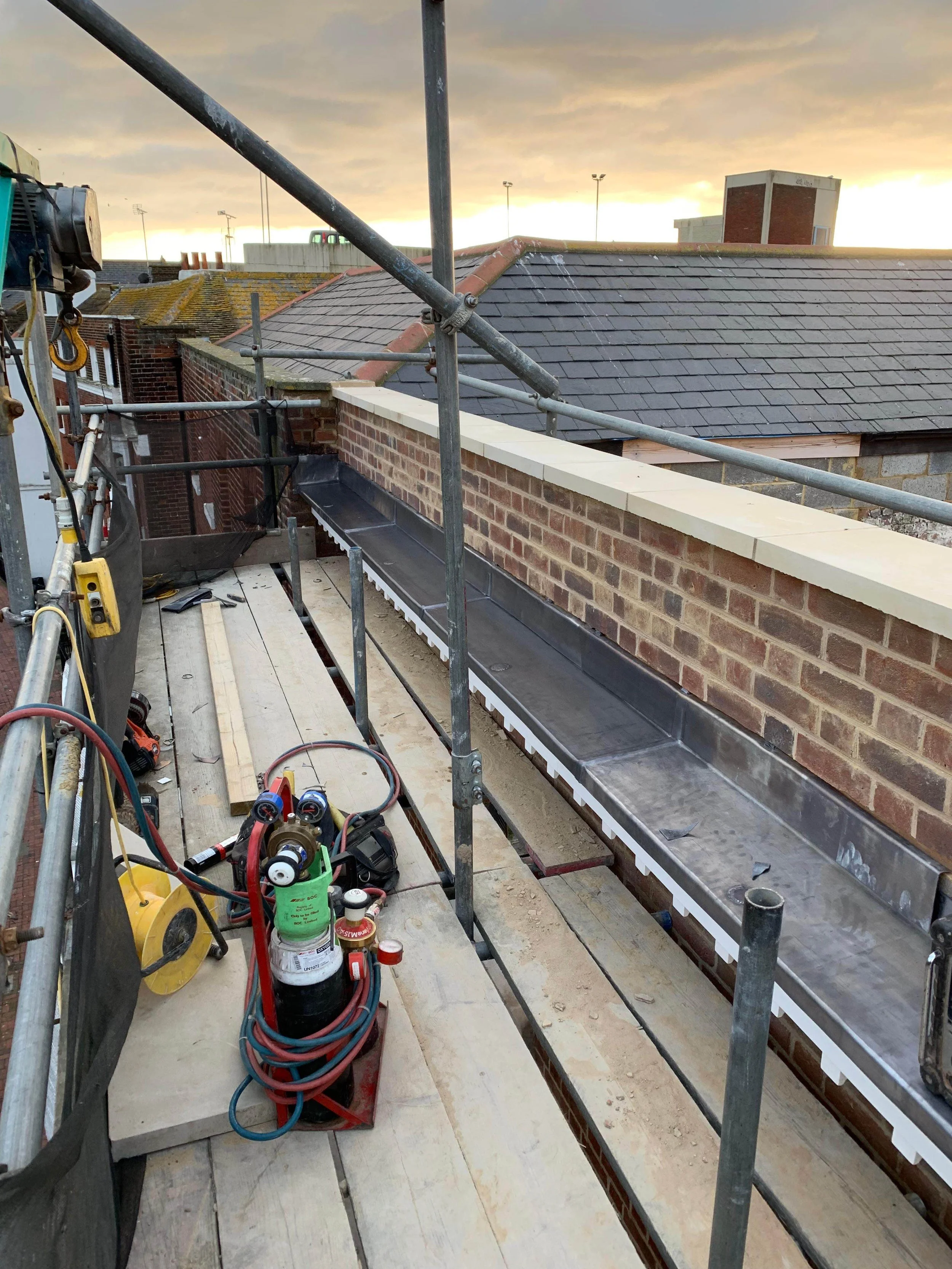 Construction scaffolding on a rooftop with tools and wiring, overlooking nearby rooftops at sunset.