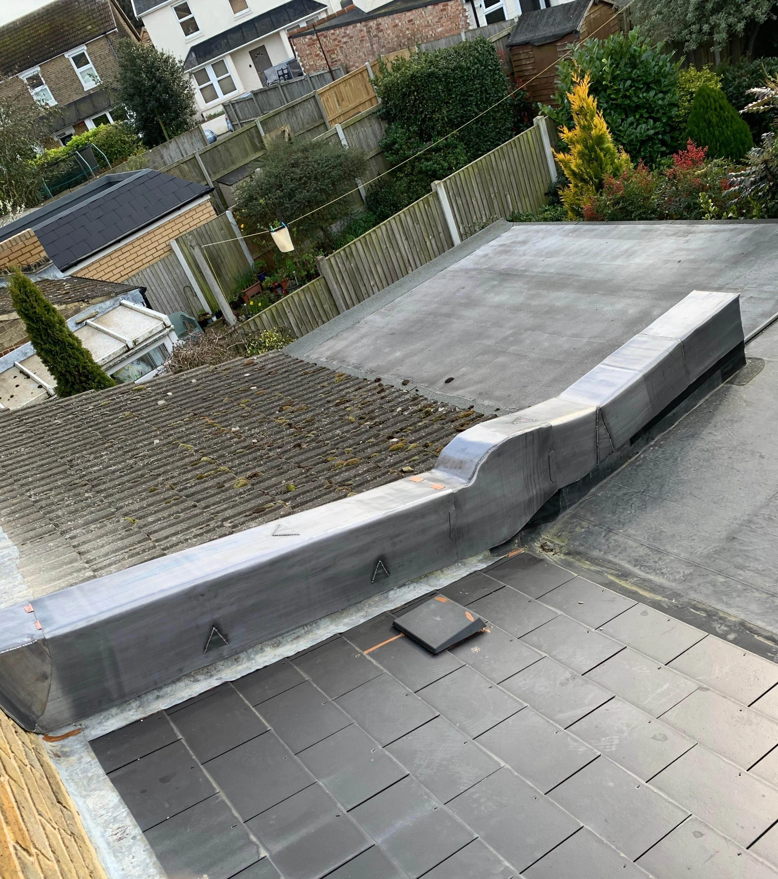 Roof with a newly installed flat roof section and a section with old, moss-covered tiles. Surrounding gardens and neighboring buildings are visible in the background.
