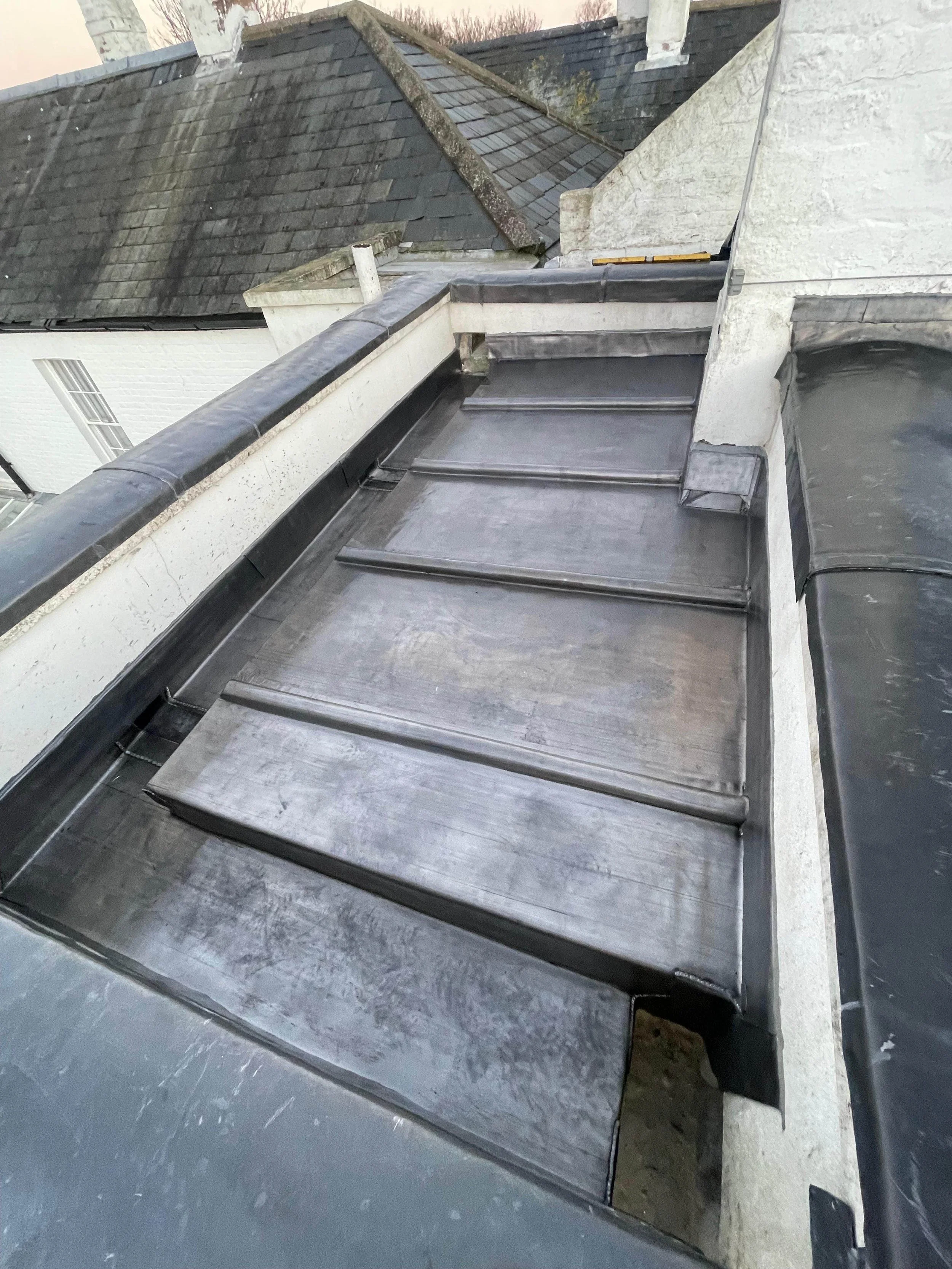 Flat roof with slate and metal roofing, part of a building's dormer window structure, surrounded by chimney and other roofing elements.