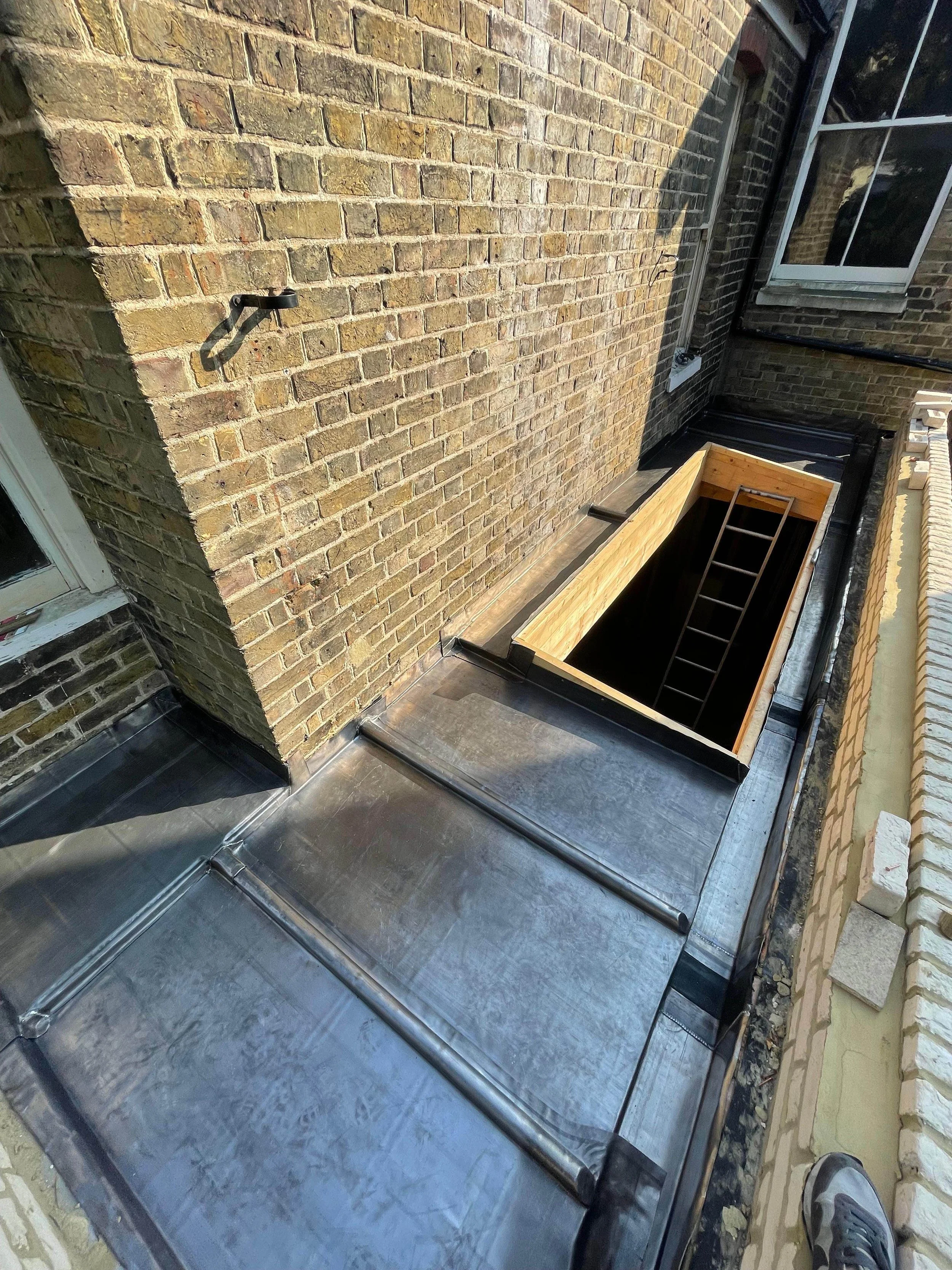View of a rooftop with metal roofing, a ladder leading into a wooden hatch, brick walls, and a window, suggesting an ongoing roofing or building renovation.
