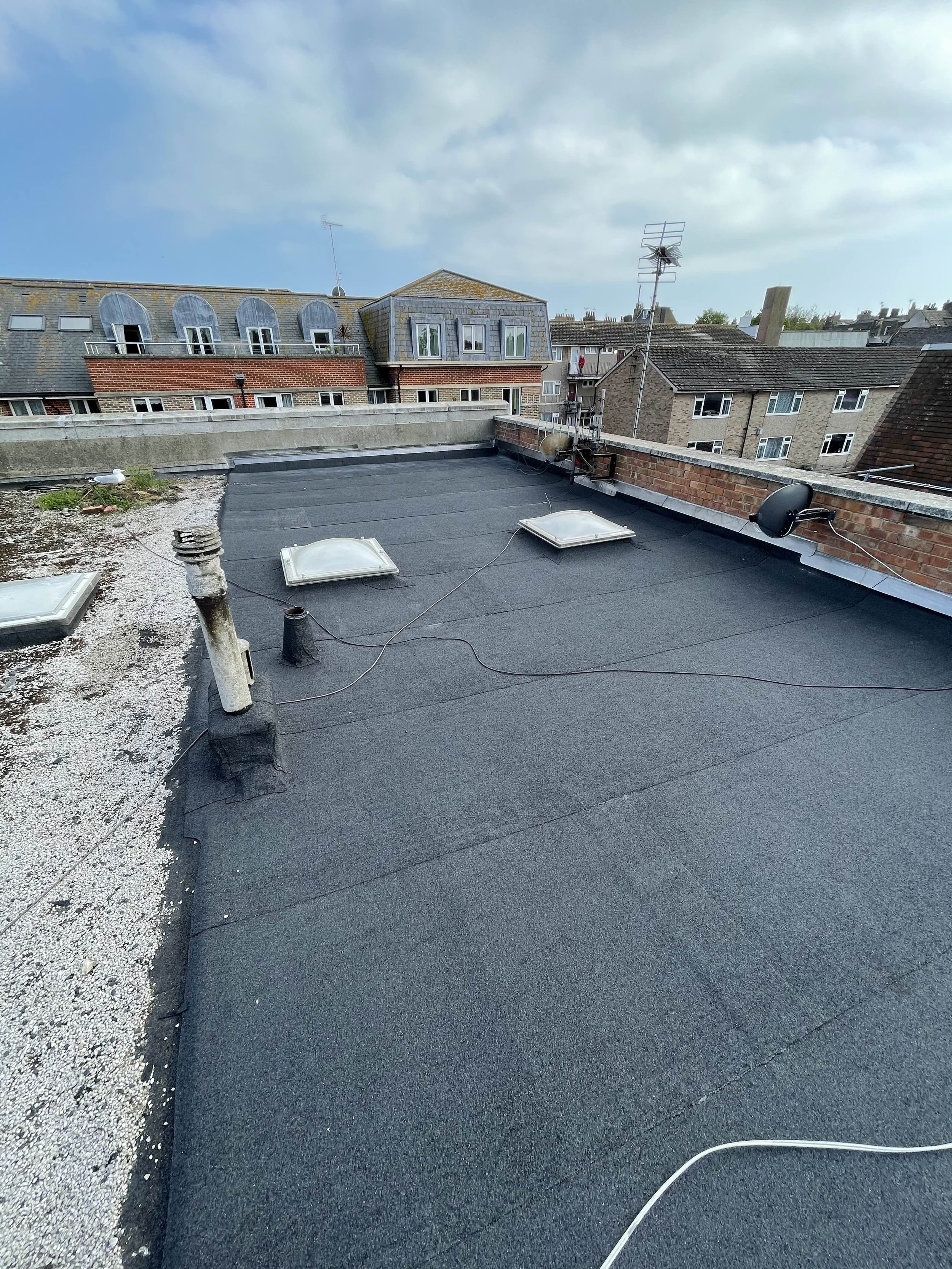 Flat rooftop with black roofing material, skylights, vents, antennas, and surrounding buildings under cloudy sky.