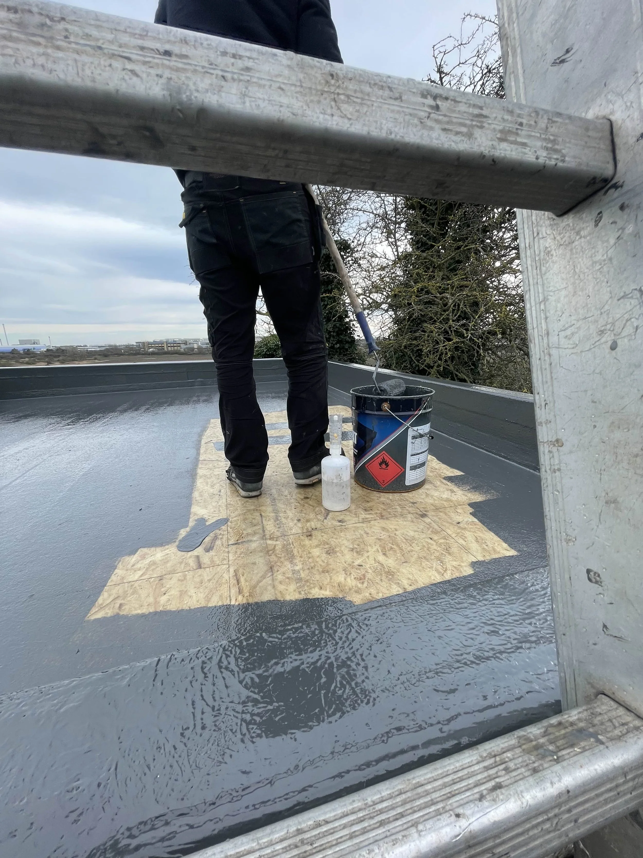 Person standing on a roof painting the surface with gray waterproof coating, seen through a ladder frame, with a bucket, a roller, and a bottle nearby.
