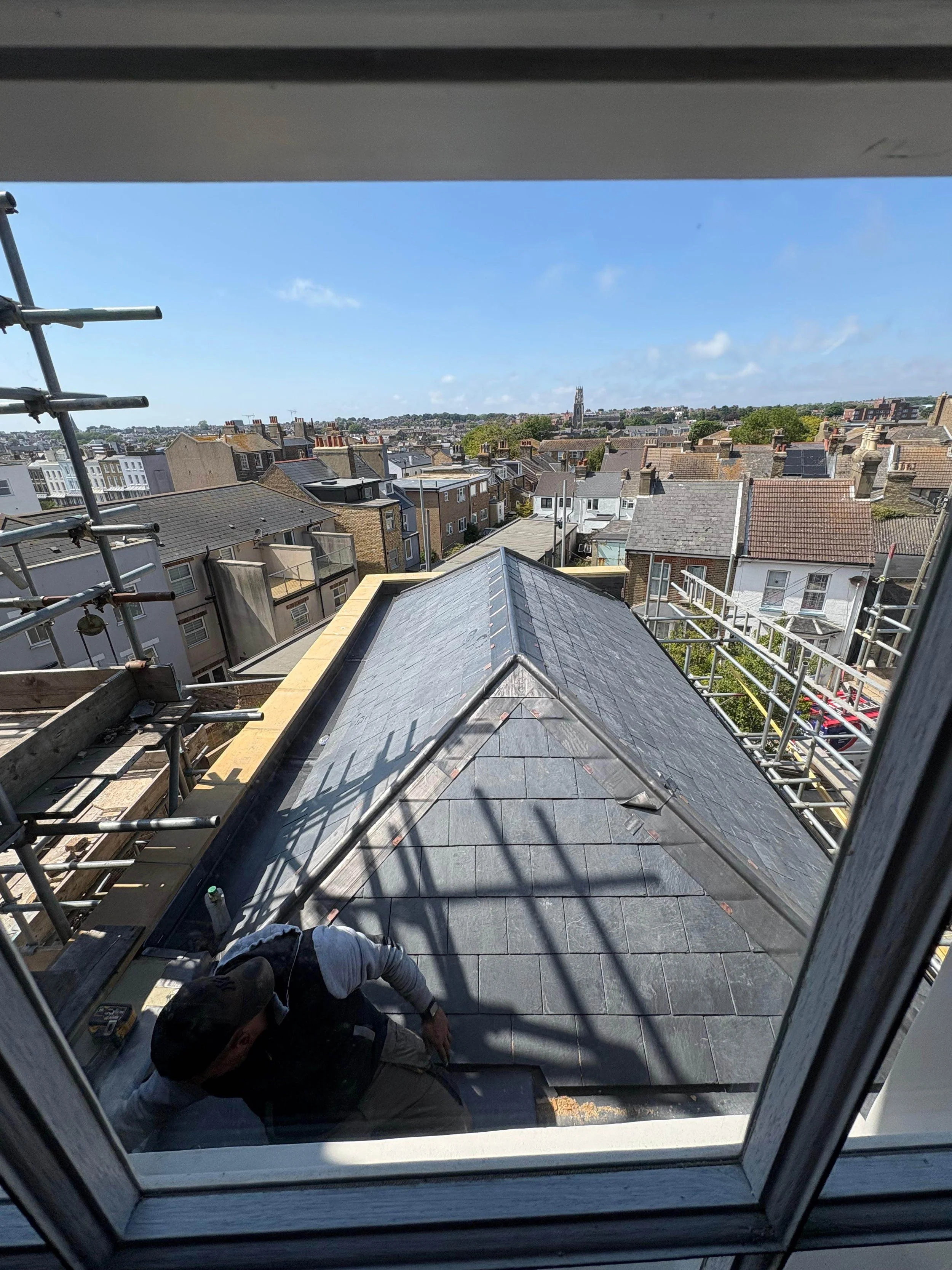 View of a newly replaced slate roof from a window, showing a person working on the roof during daytime with a residential neighborhood and blue sky in the background.