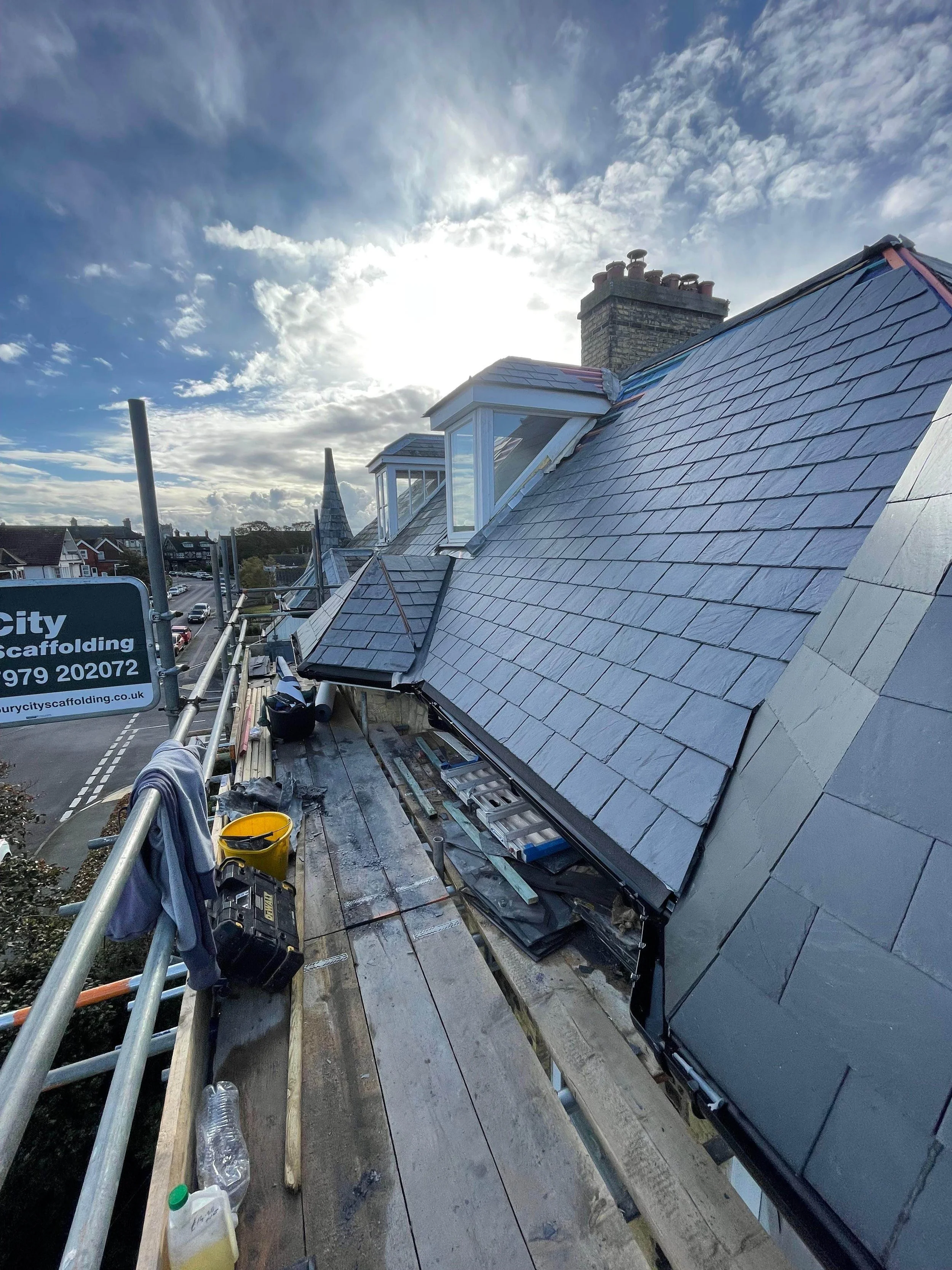 Construction scaffolding on a rooftop with roofing materials and tools, a sign for City Scaffolding, and a view of neighboring buildings against a partly cloudy sky with the sun shining.