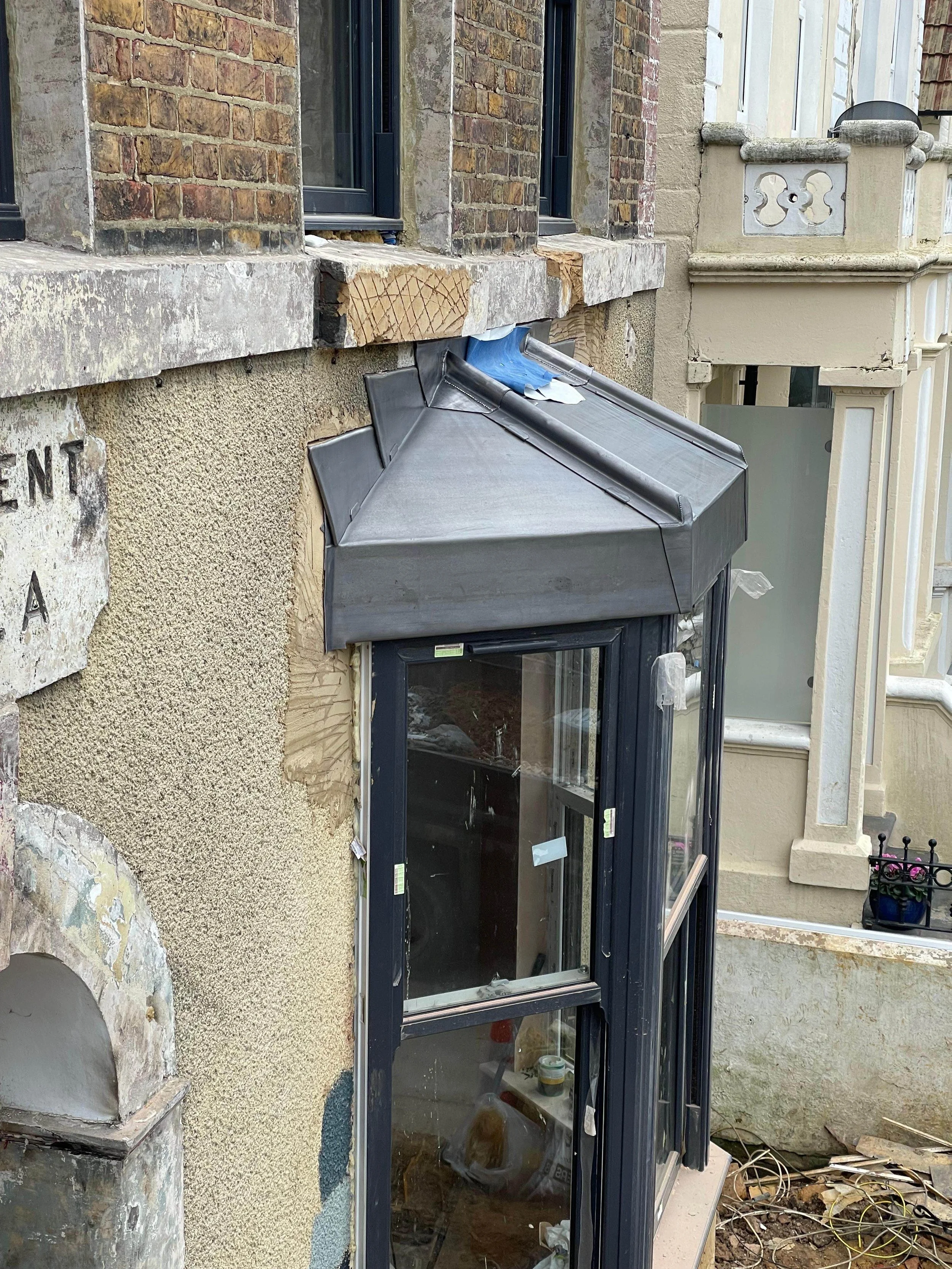 A close-up of a building exterior showing a window with a black metal frame and transparent glass, a small black metal awning above the window, and construction or renovation work with exposed materials and debris nearby.