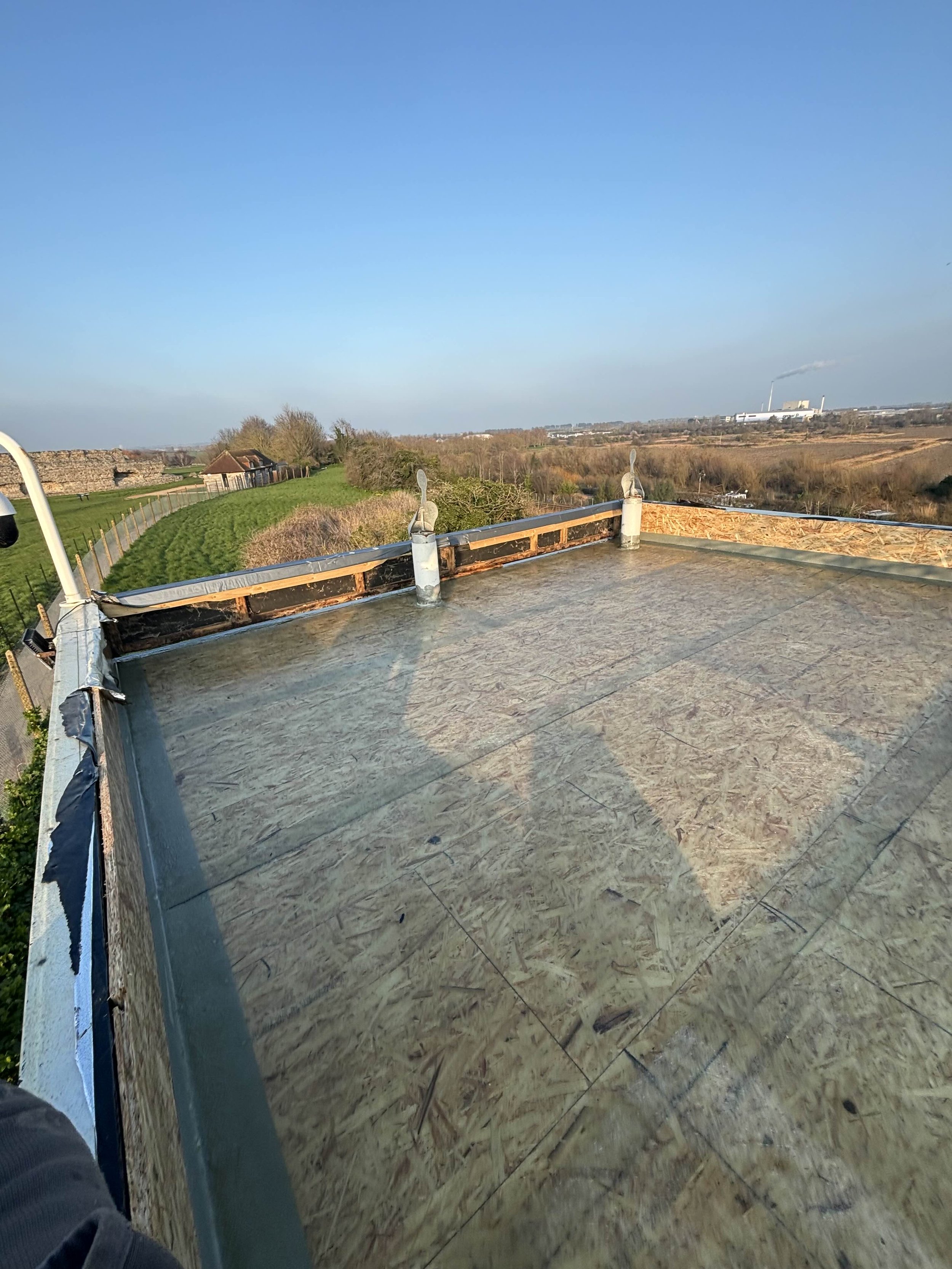 View from the roof of a building under construction with a flat surface covered with oriented strand board and orange foam insulation. The roof has some metal vents and is surrounded by a metal edge. In the background, there are open fields, trees, a