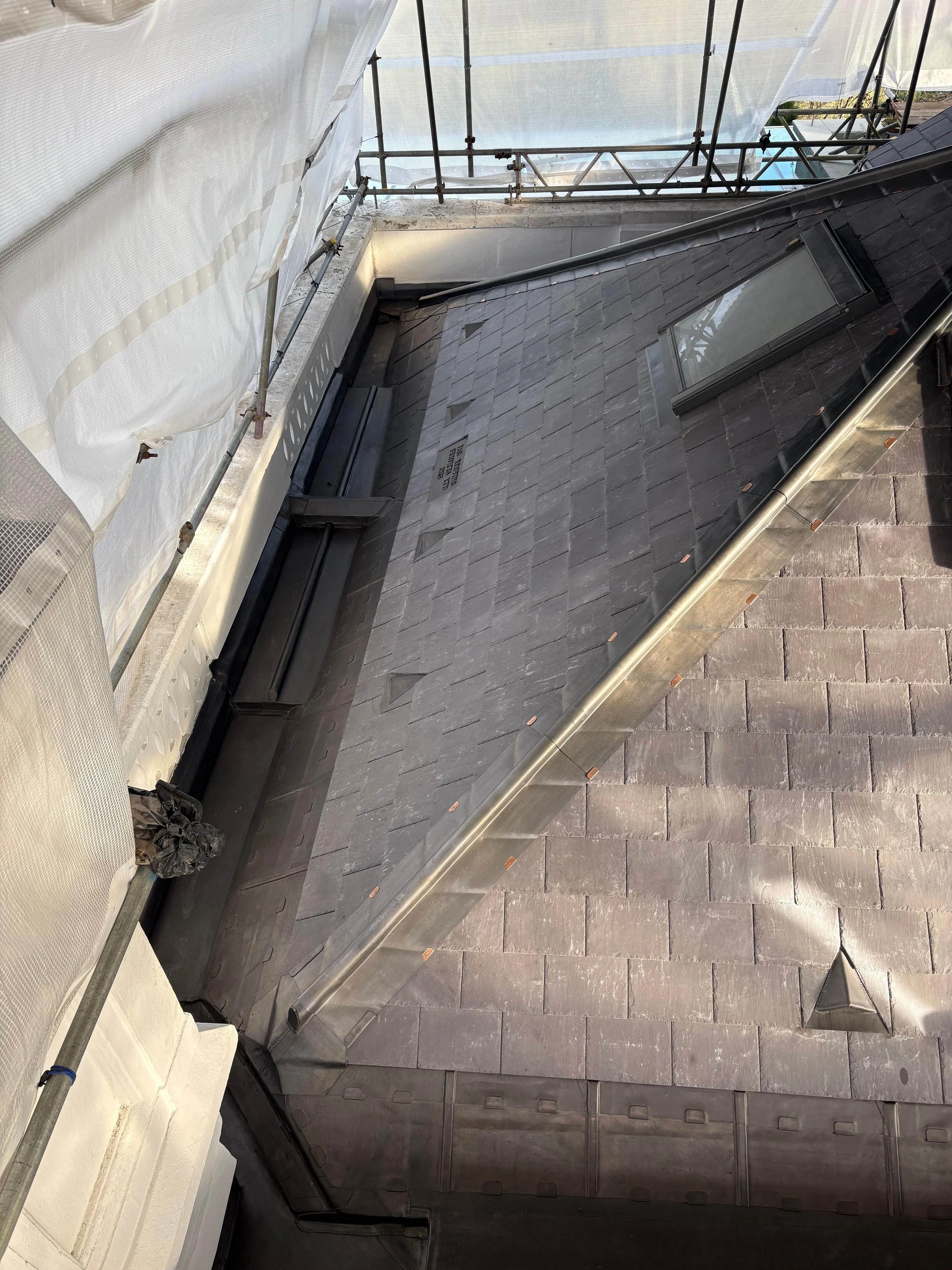 A roof with slate tiles under construction, showing a skylight window and a metal vent pipe, with scaffolding and white construction wrap on the side.