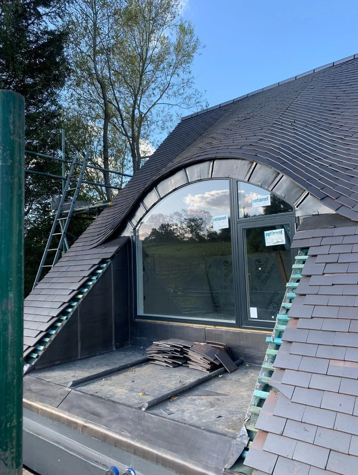 Under construction rooftop with a large arched window, roofing materials, scaffolding on the side, and a clear sky with some clouds in the background.