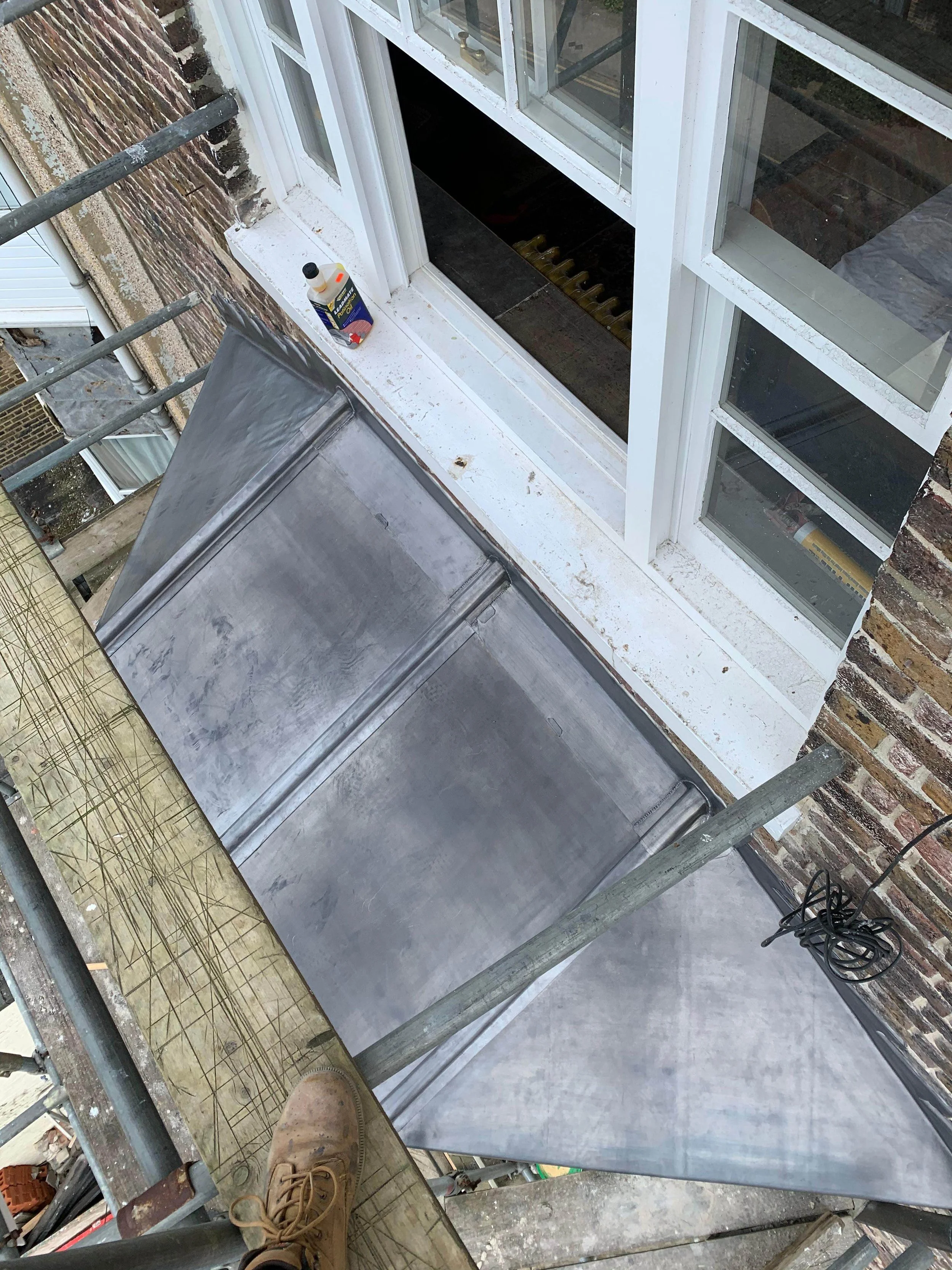 View from scaffolding showing a partially installed metal roof on a building with a white-framed window and brick wall.