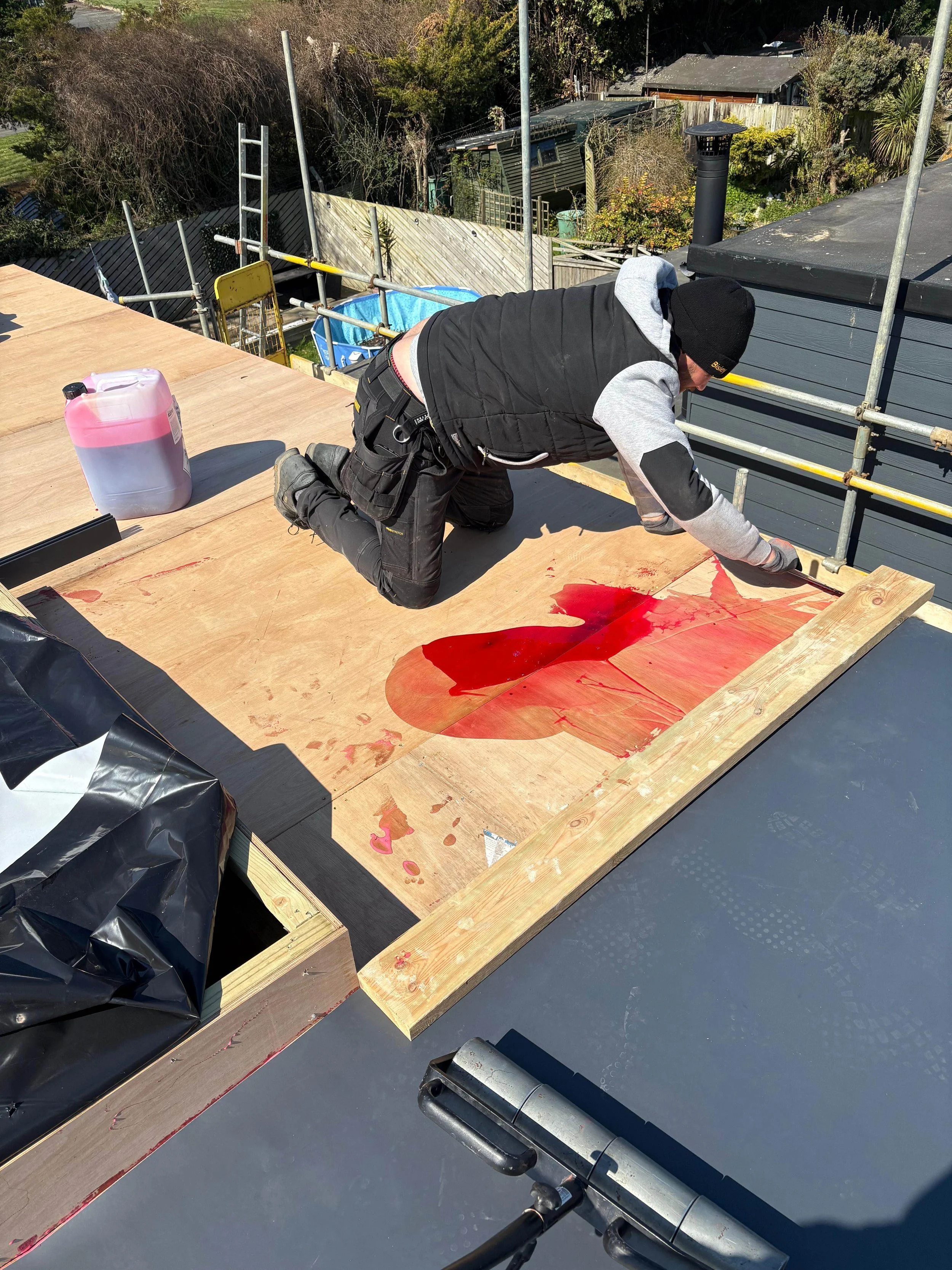 A construction worker kneels on a wooden roof, applying red roofing adhesive or sealant. The worker is dressed in a black vest, gray sleeves, black pants, and a black beanie. There are tools and a container of red liquid nearby on the roof.