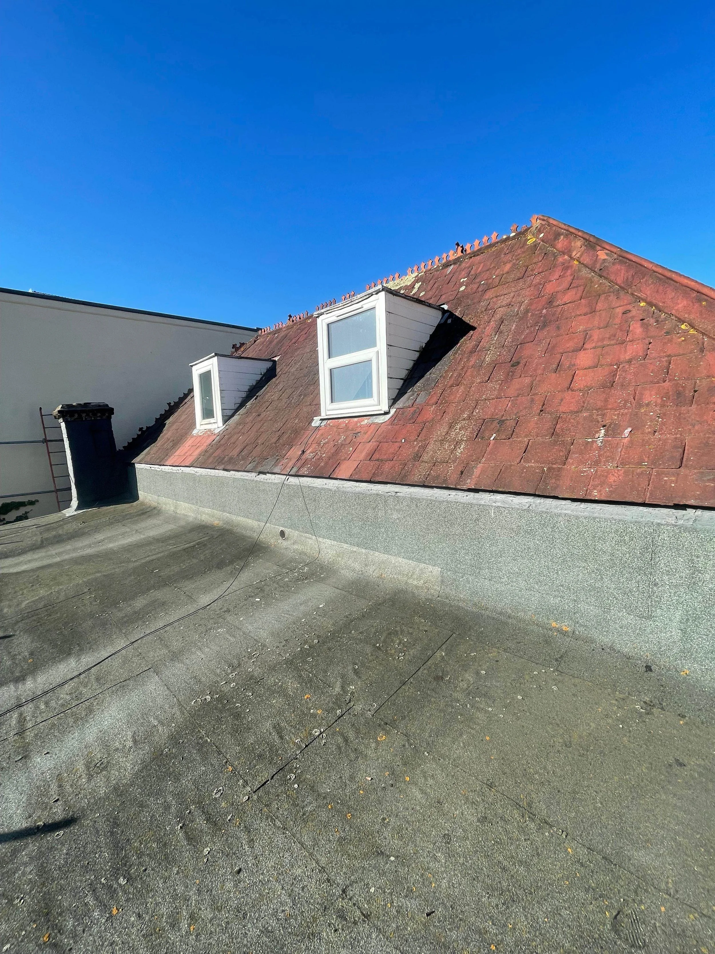 View of a rooftop with two dormer windows, red shingles, a chimney, and a flat section with a weathered surface under a clear blue sky.