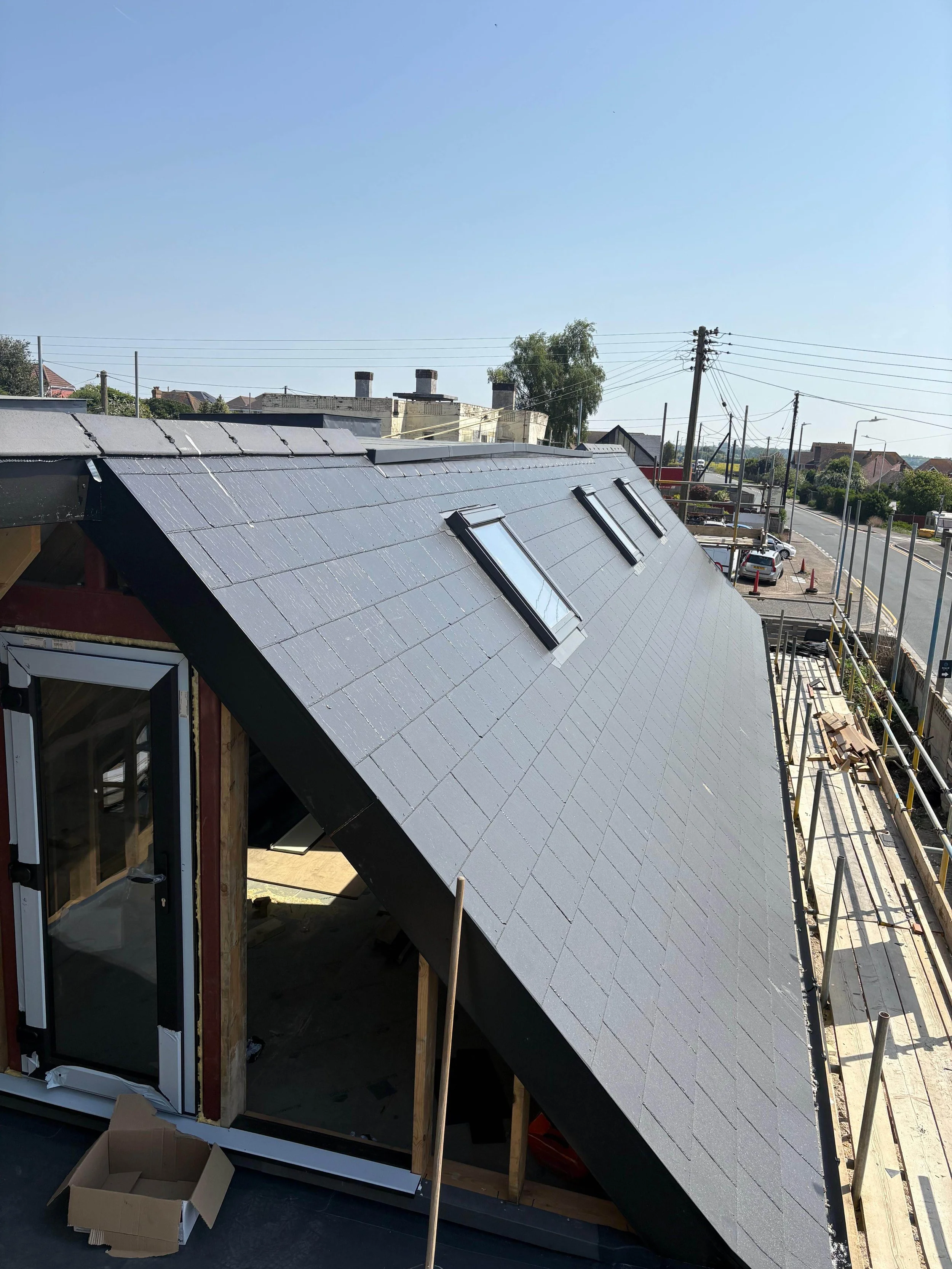 Construction site with a new roof, skylights, and scaffolding on a building, with a view of a street, parked cars, and power lines in the background.