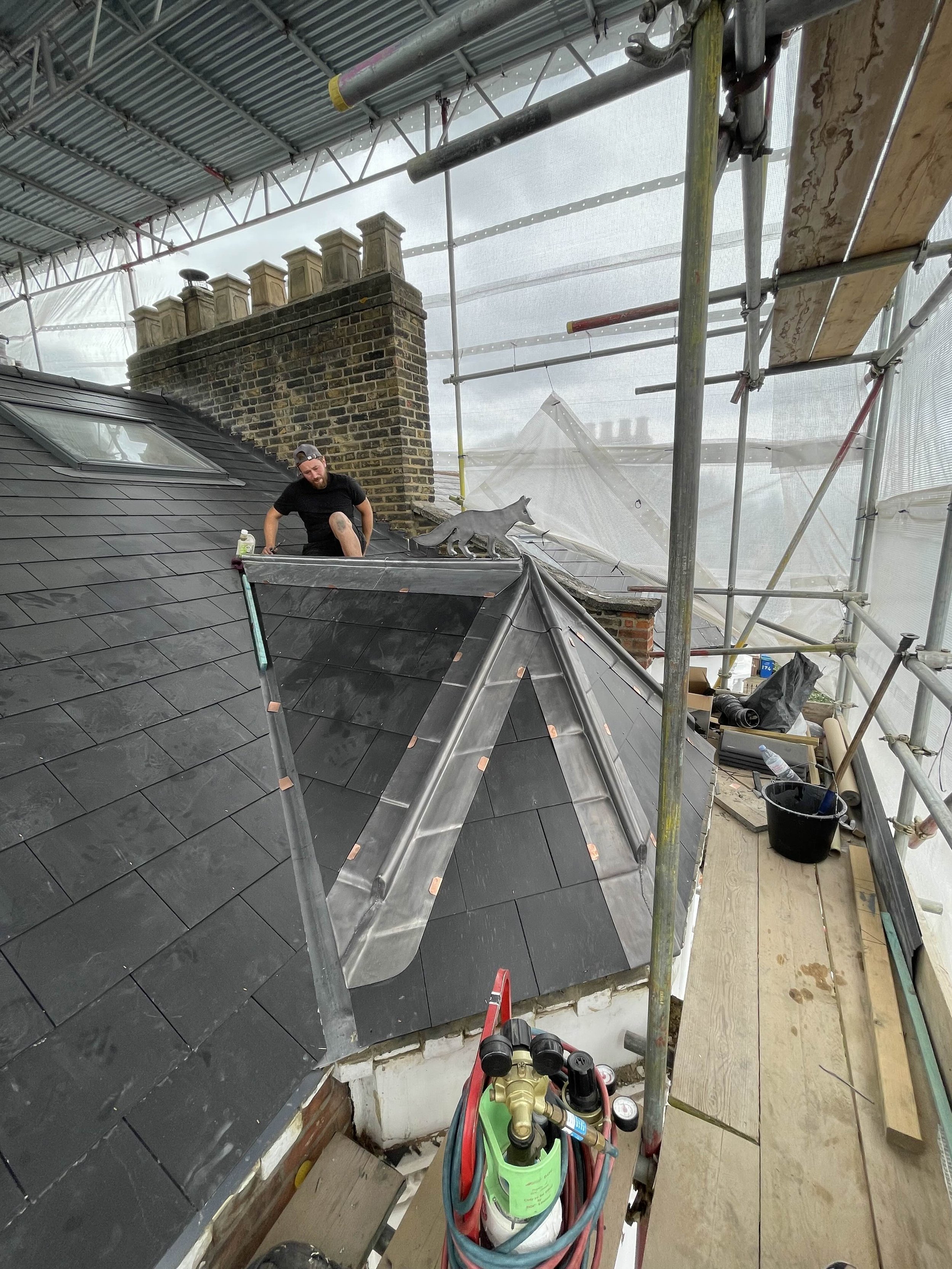 Construction worker on a roof installing slate shingles, surrounded by scaffolding and construction tools, with a brick chimney in the background.