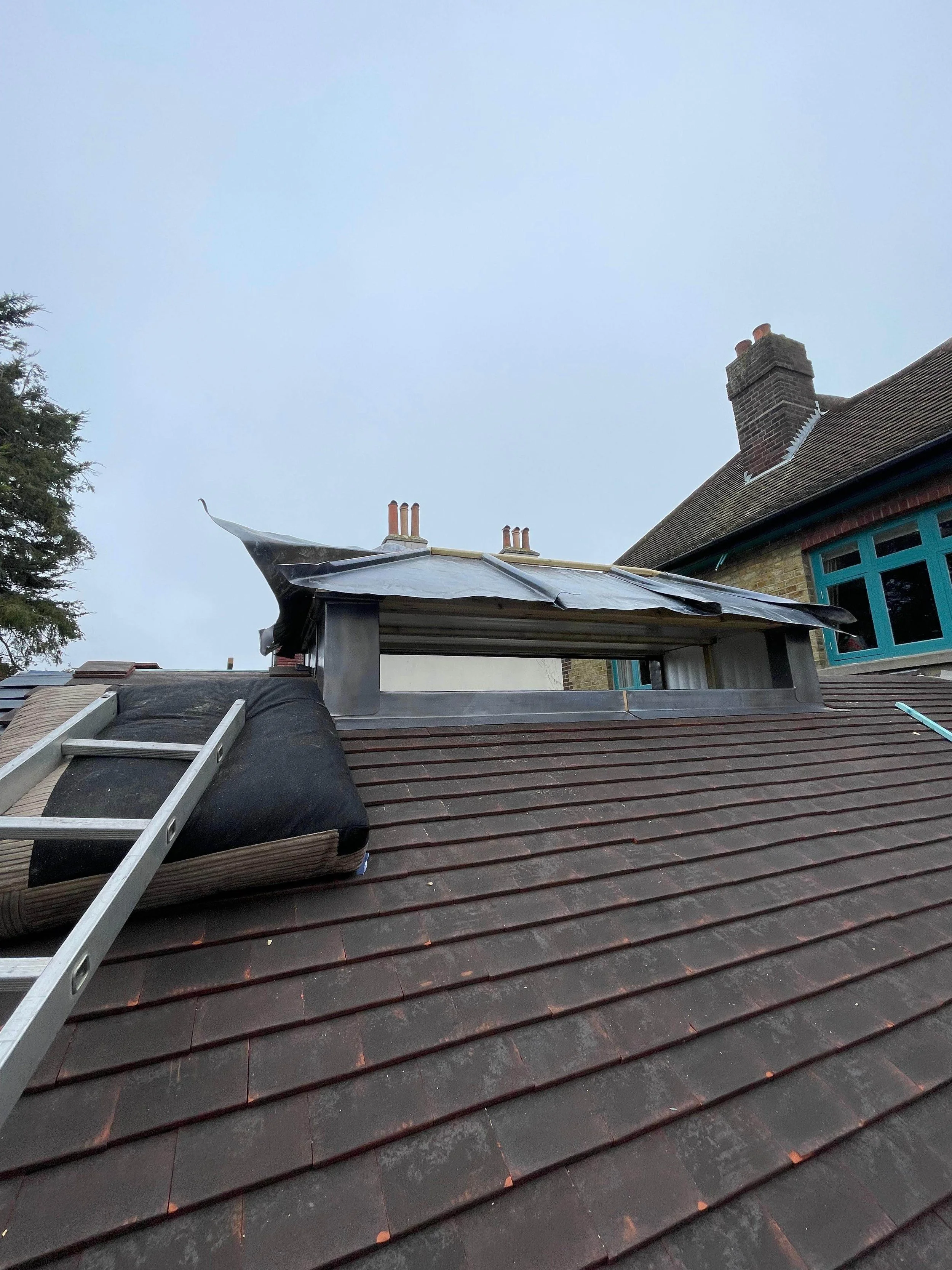 Roof with ladder, insulation, and metal flashing around a chimney and roof window, overcast sky.