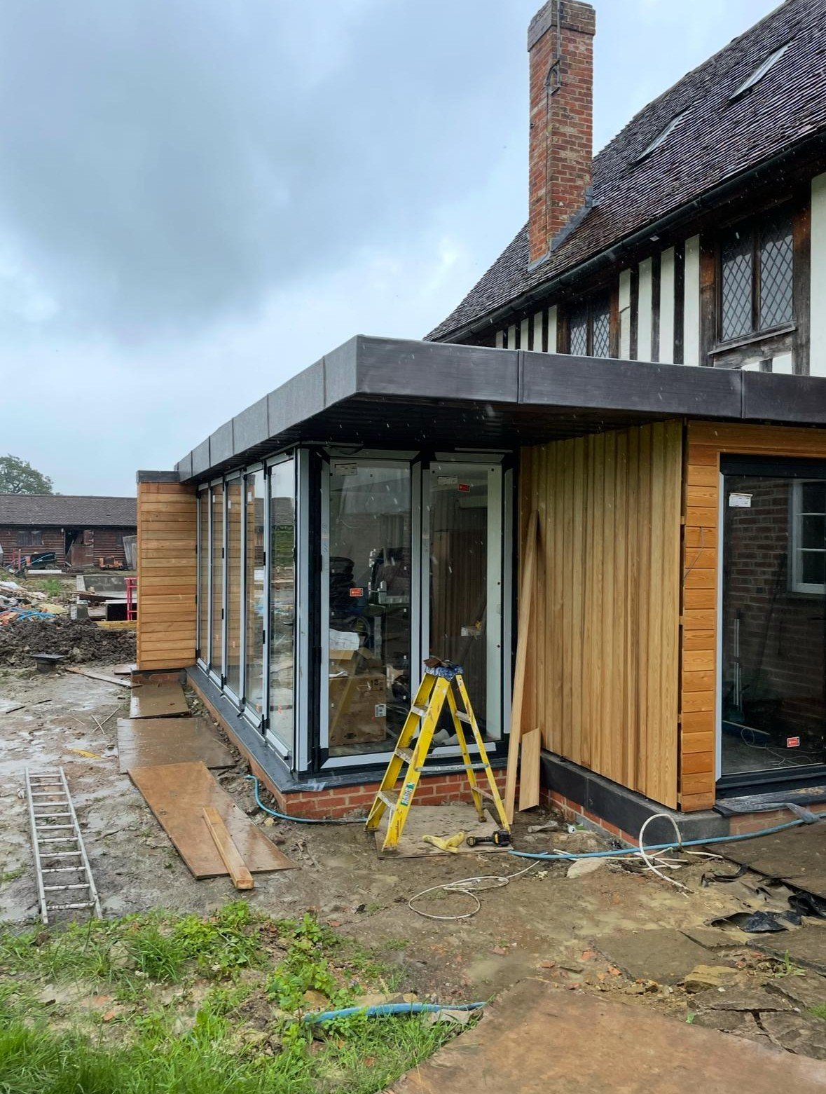 Construction site of a house extension with glass walls and wooden siding, tools and equipment in the foreground.