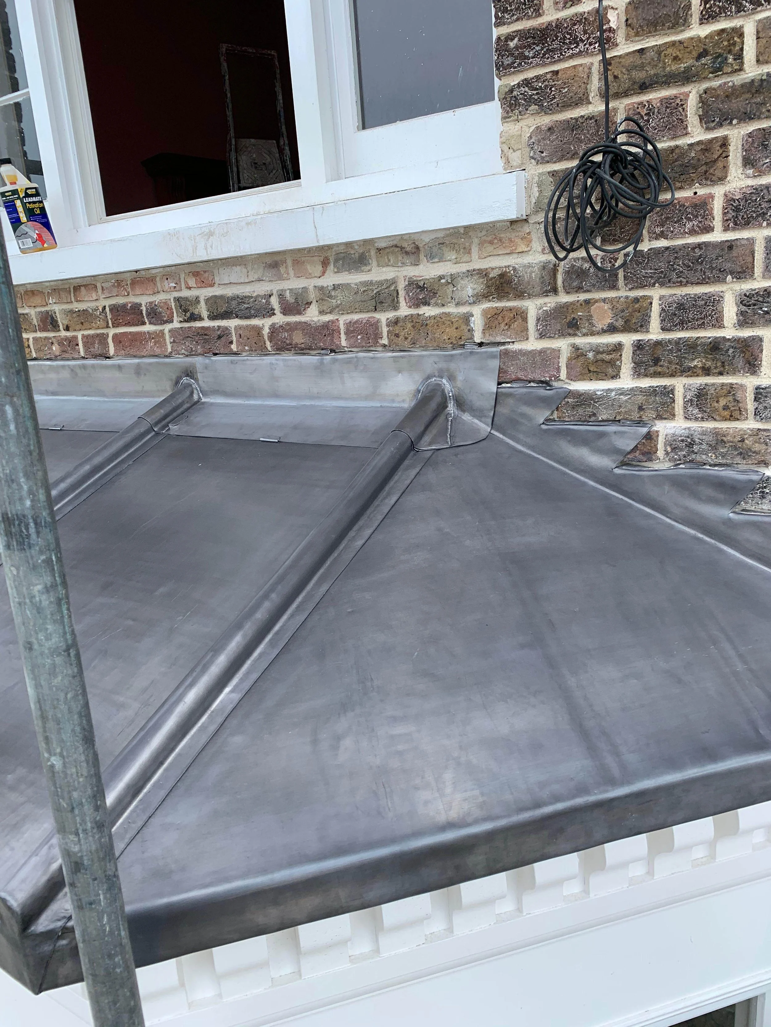 Close-up of a metal flashing process on a brick wall exterior near a white window. The metal is freshly installed, with corner bends and seams, and an electrical cord is coiled on the brick wall nearby.
