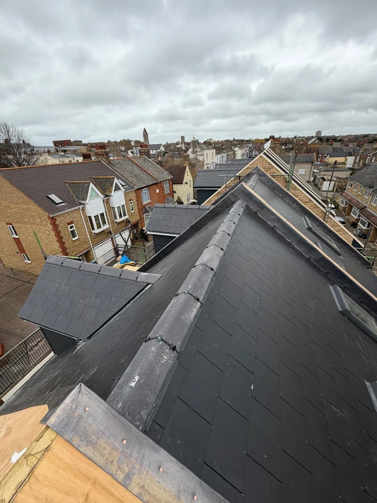 View from a rooftop showing surrounding houses with black and grey roofing, overcast sky, and a distant church steeple.