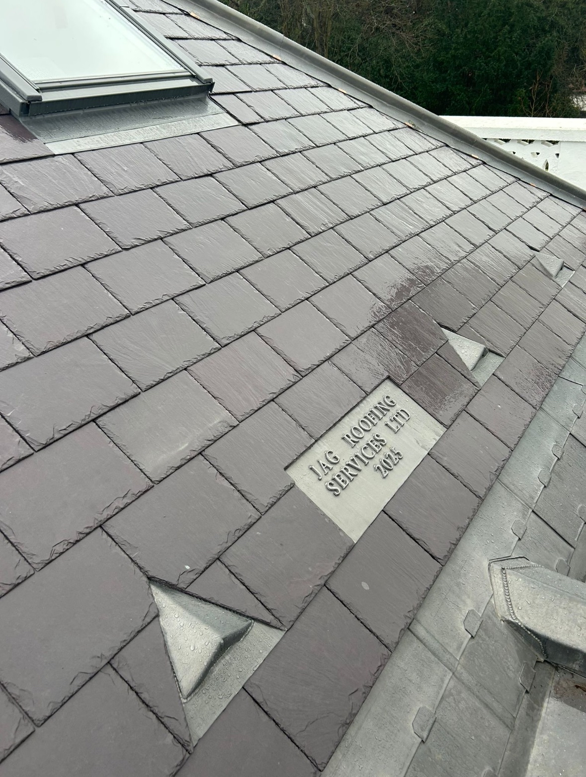 Close-up view of a gray tiled roof with a metal plaque that reads 'JAG ROOFING SERVICES LTD 2015'.