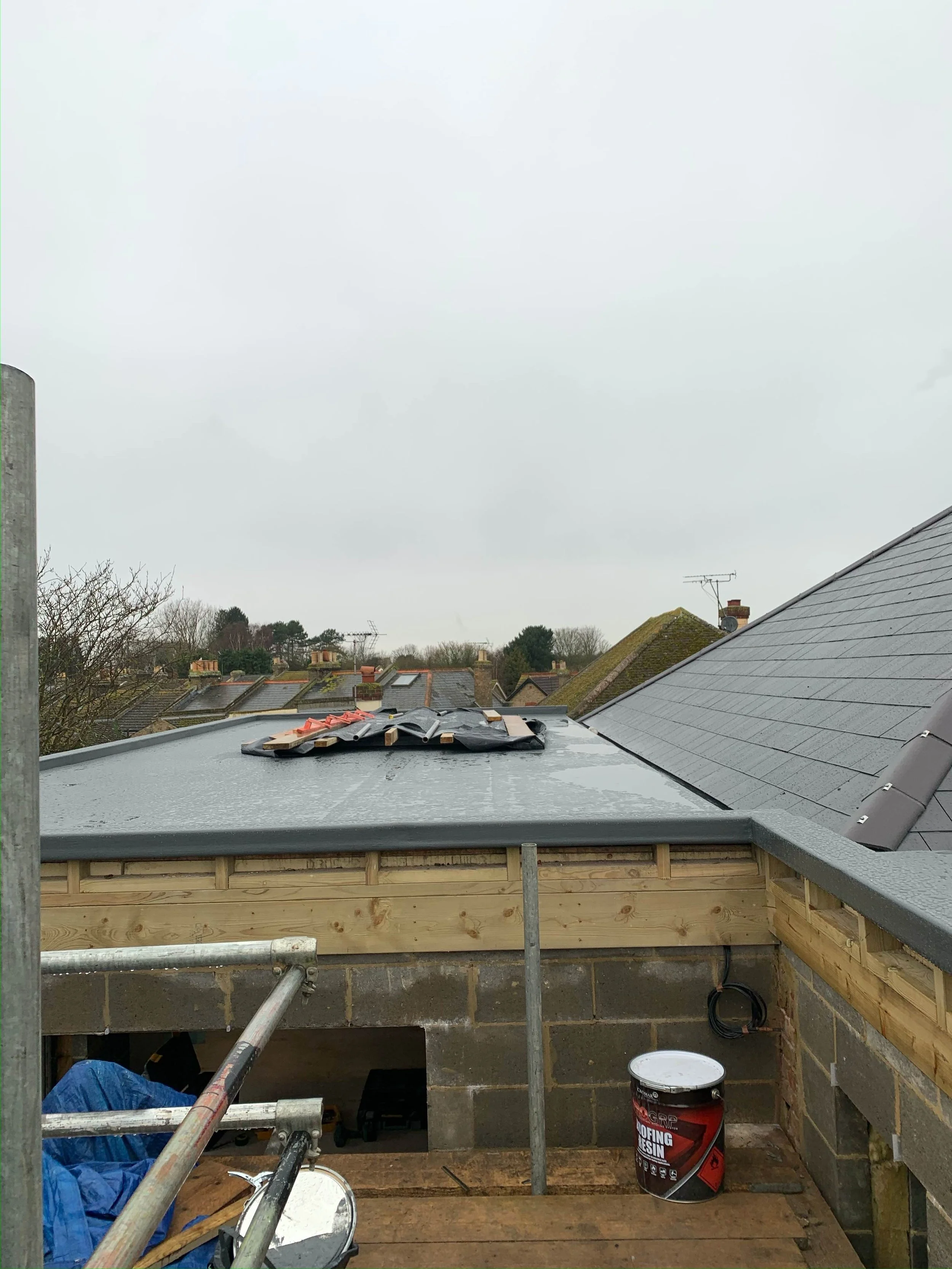 Roof construction site with roofing materials, scaffolding, and a bucket of roofing resin against an overcast sky.