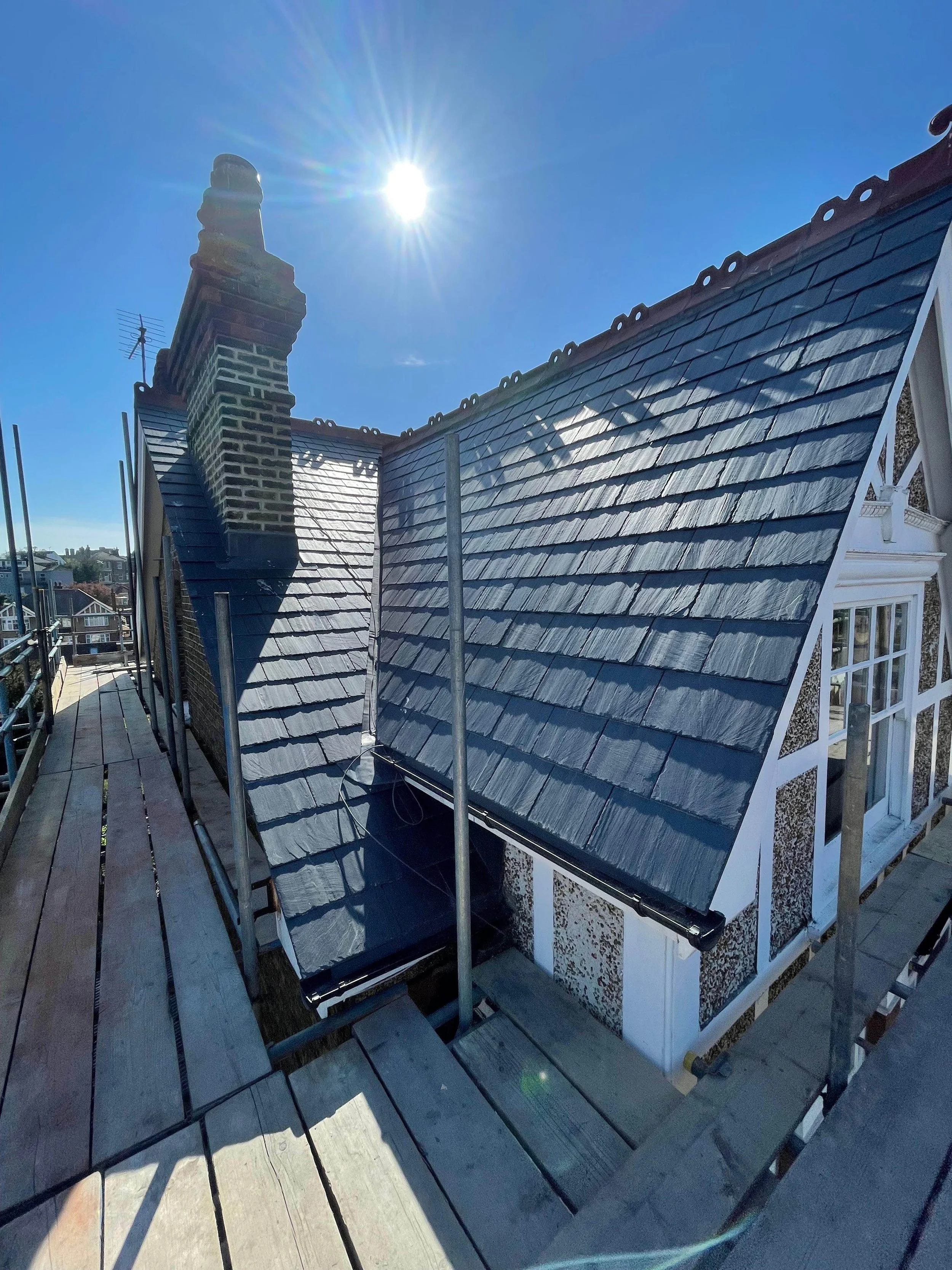 The image shows a rooftop with slate shingles, scaffolding, and a chimney with smoke stack. The sky is blue with the sun shining brightly overhead.