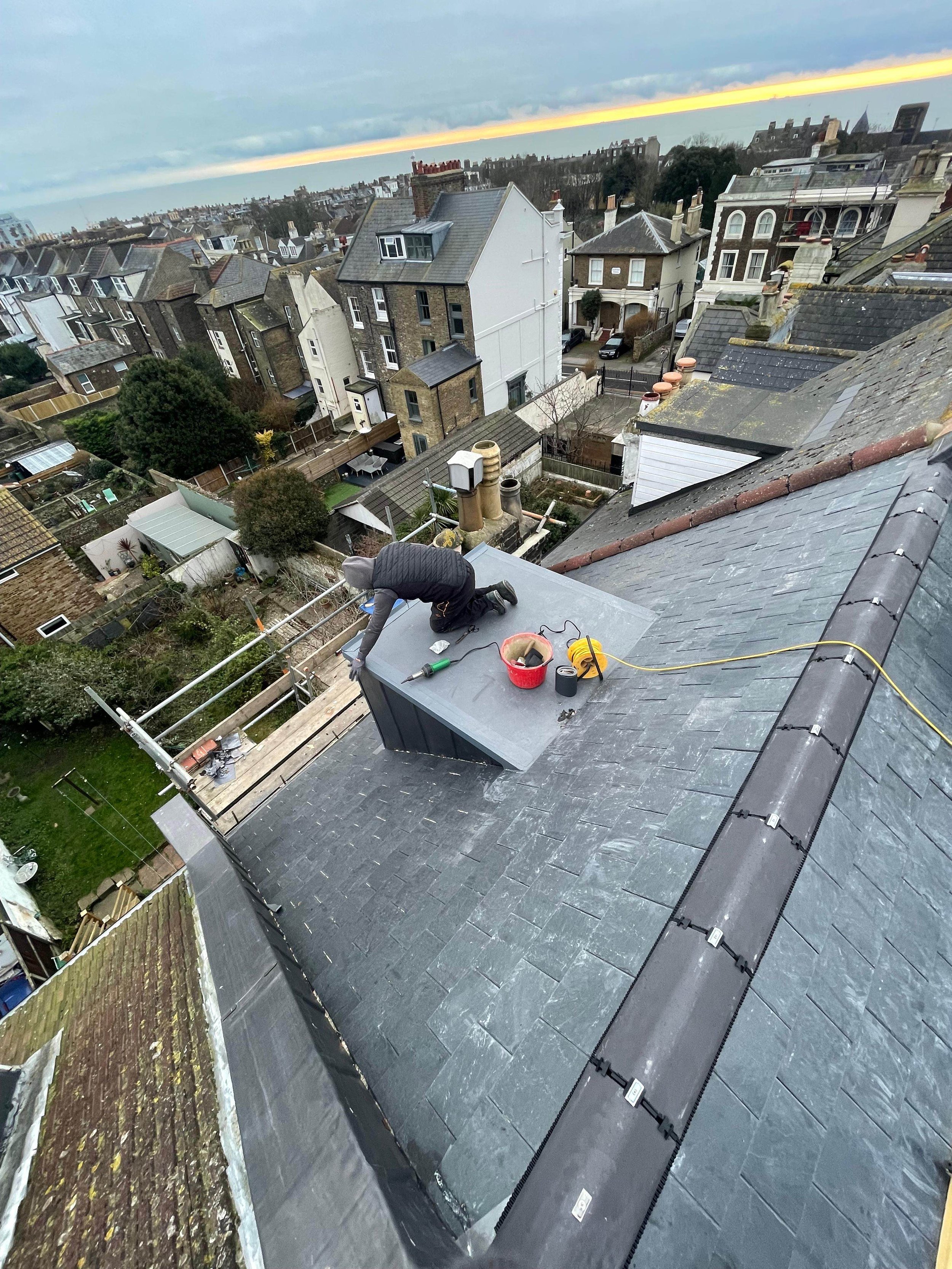 A person working on a rooftop with tools, surrounded by neighboring rooftops and houses during dusk with a colorful sunset in the background.