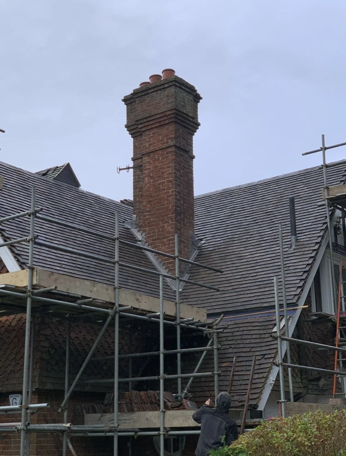 A house with a brick chimney undergoing roof repairs, scaffolding surrounds the house, and a person is pointing towards the roof.