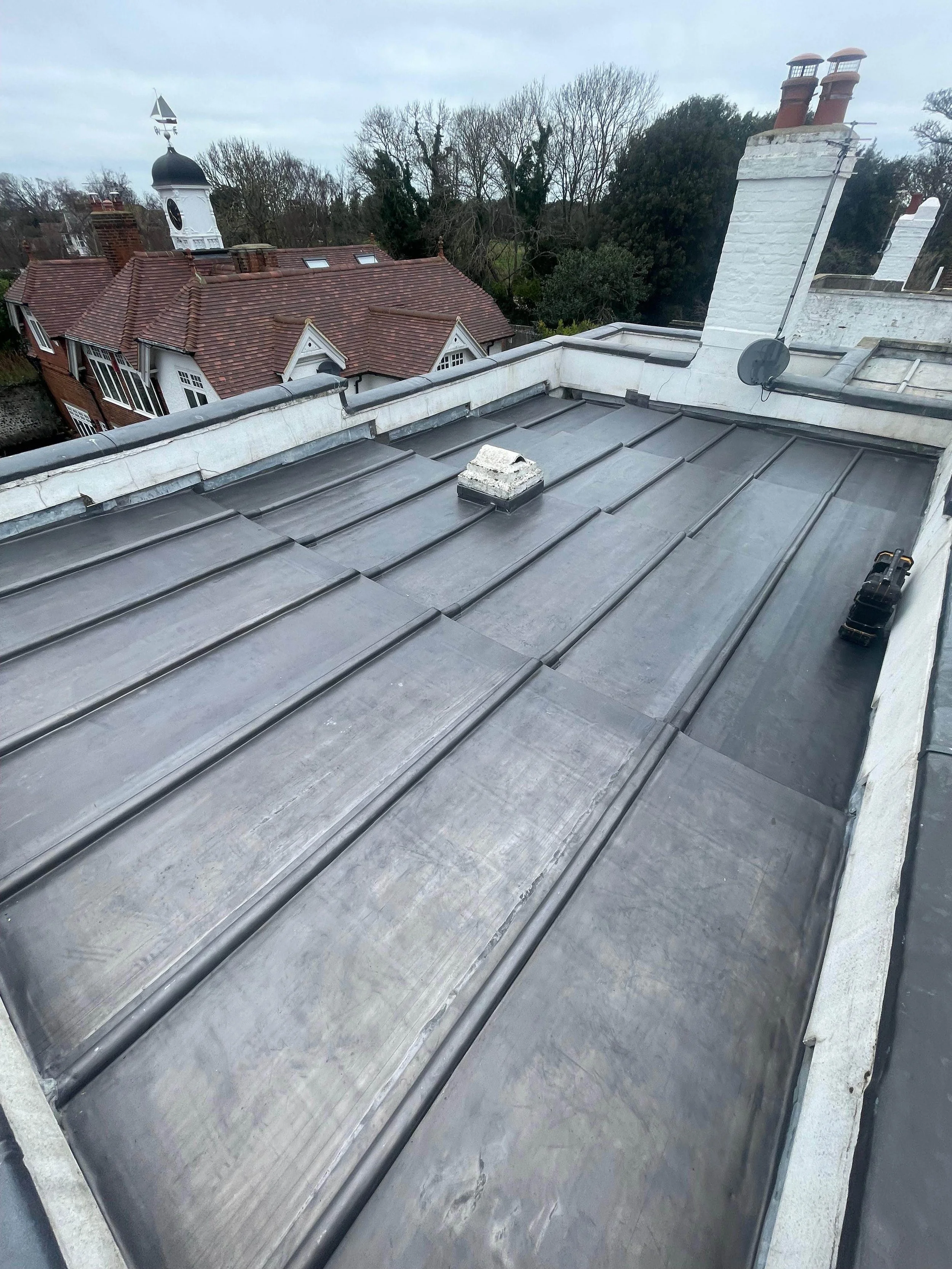 View of a metal roof on a building with surrounding chimney stacks, chimney pots, trees, and neighboring houses with tiled roofs and a clock tower in the background.