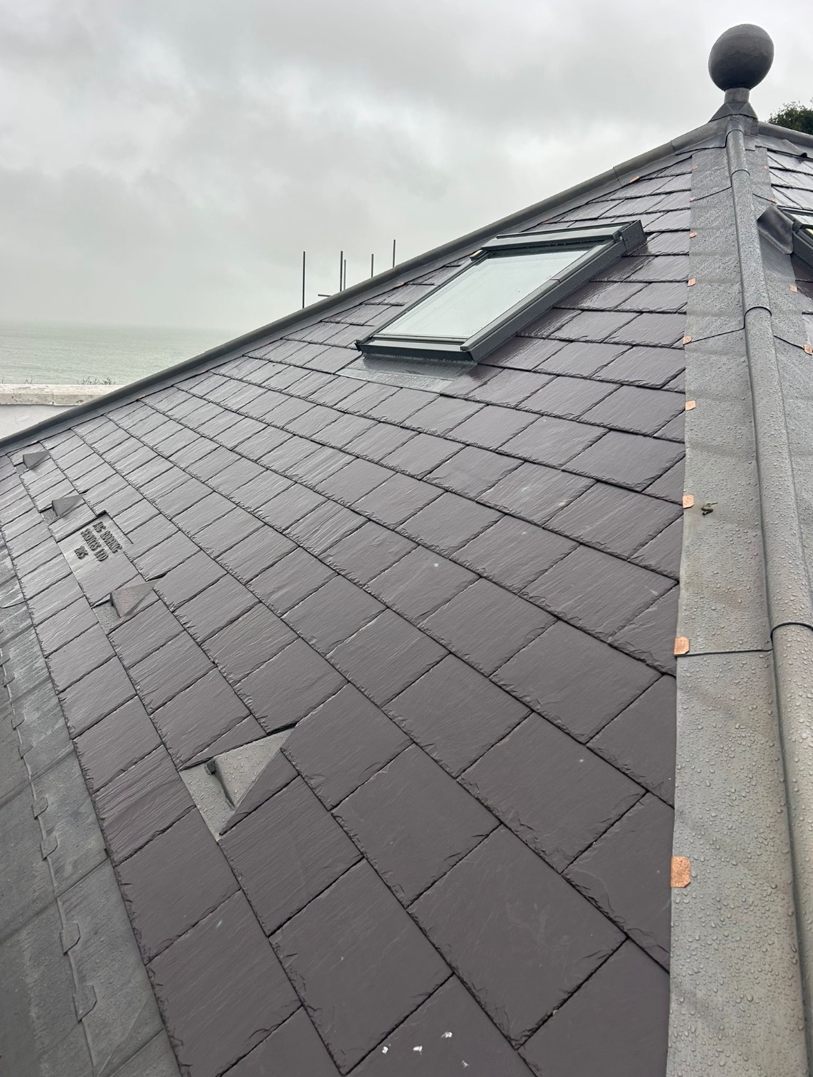 Close-up of a gray tiled roof with a skylight window and a cloudy sky in the background.