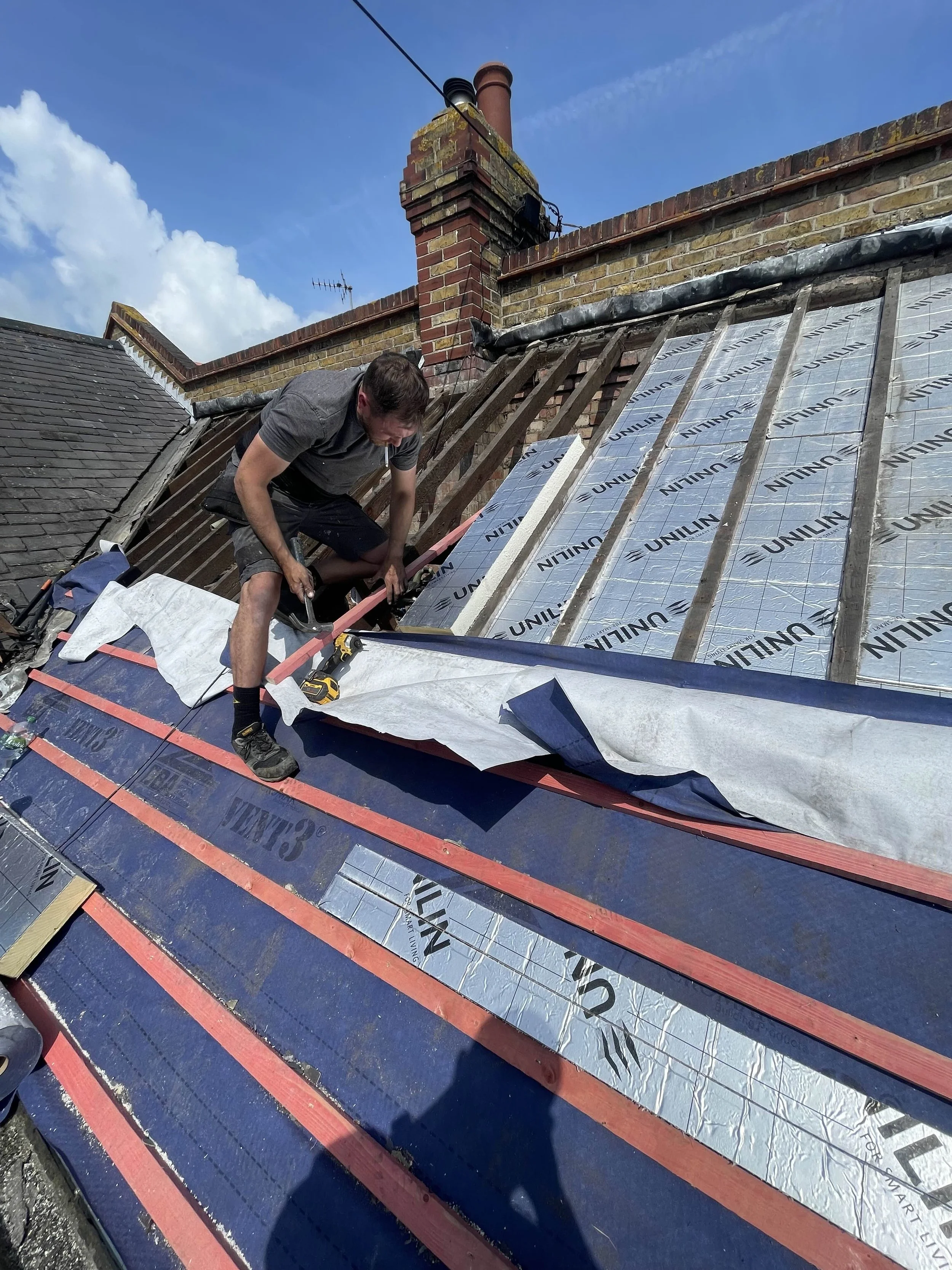 A man working on a roof, installing insulation and underlayment, with a partly cloudy sky in the background.