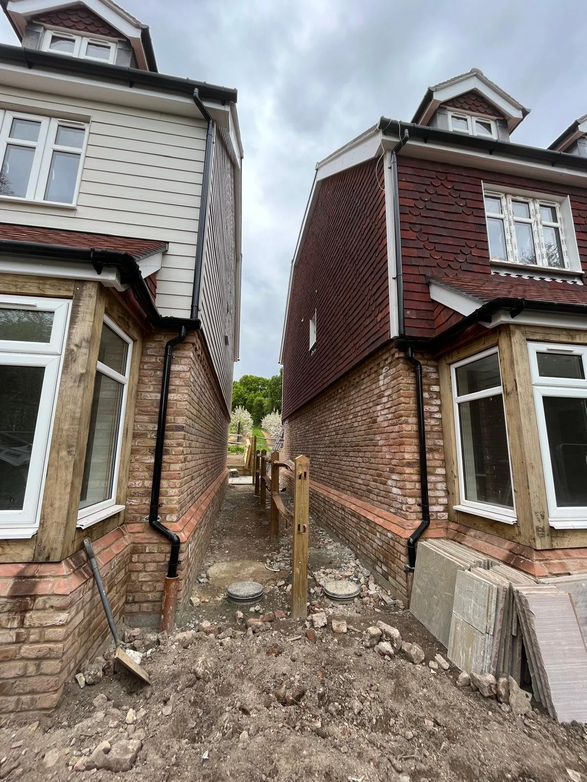 Two newly constructed houses with brick basements and varying siding on the upper floors, separated by a narrow pathway under construction, with construction materials and tools on the ground.